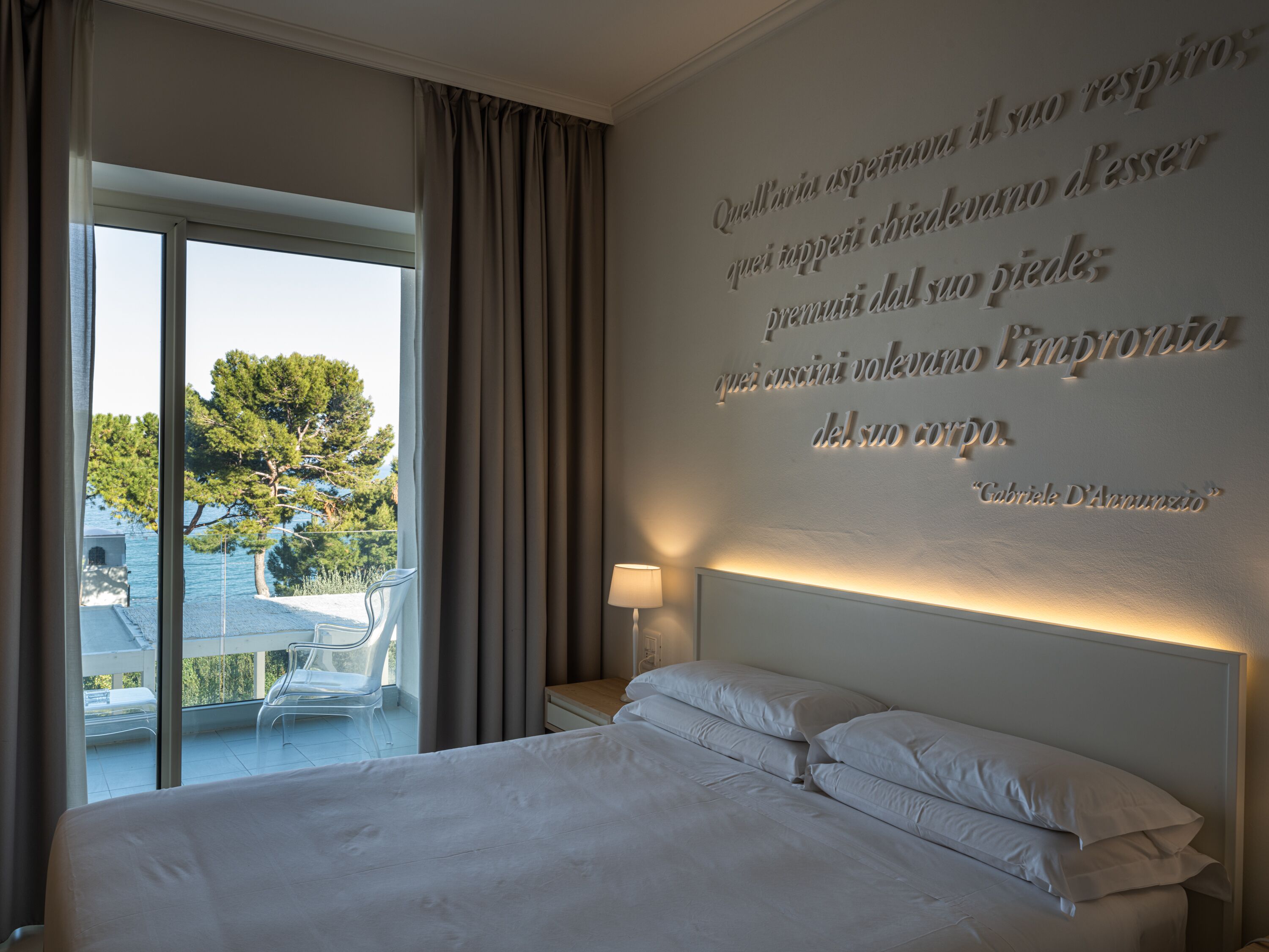 Una camera d'hotel con un letto grande, un comodino, una lampada e vista all'esterno attraverso un'ampia finestra.