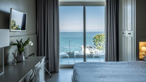 A hotel room with a bed, a desk with a chair, a vase with flowers, a TV on the wall, and a view of the sea through a large window. - Hotel La Chiave dei Trabocchi (San Vito Chietino)