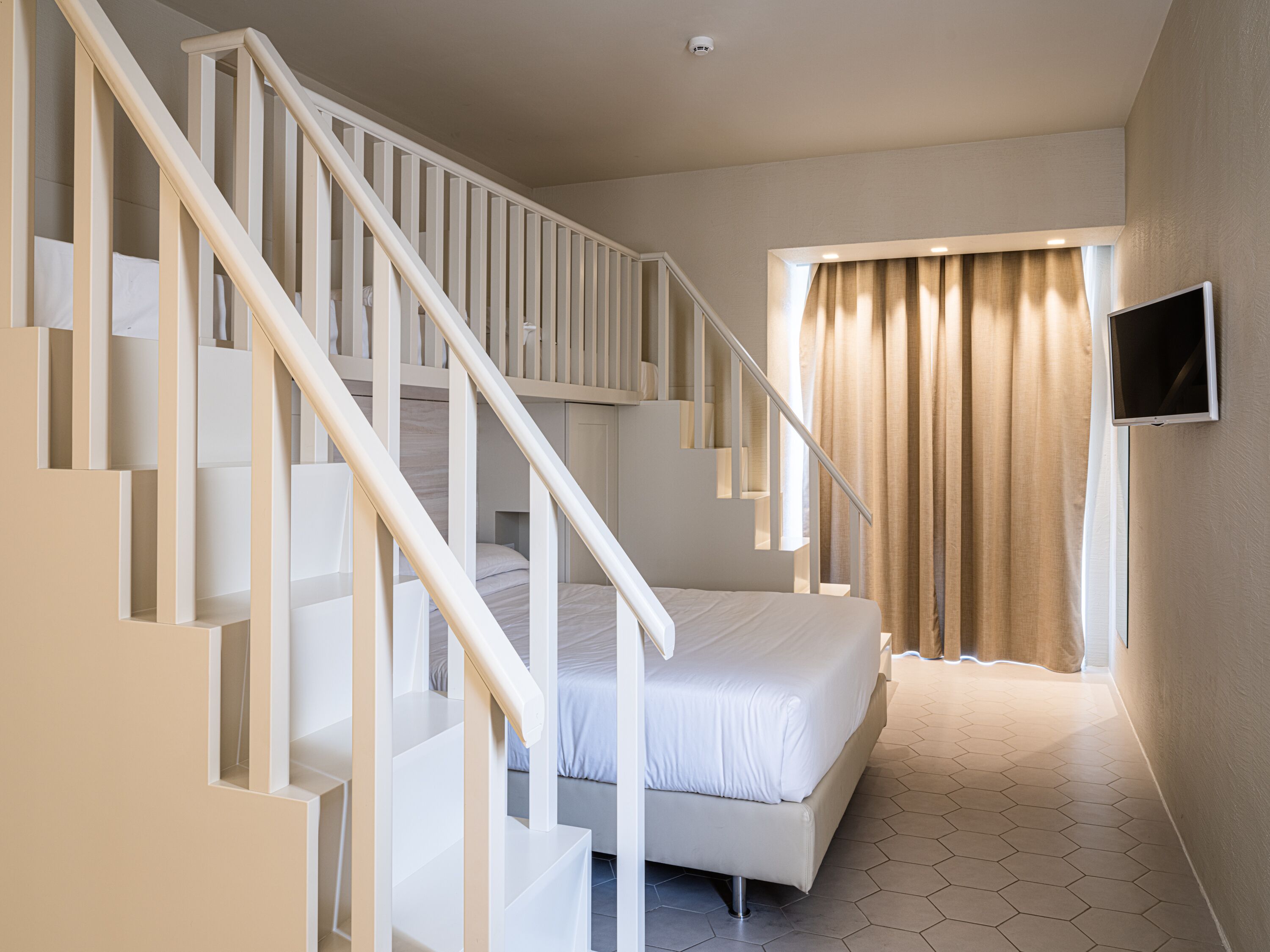 Un dormitorio con cama blanca, una escalera, un televisor montado en la pared y una ventana con cortinas.
