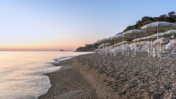 A pebbled beach with umbrellas and lounge chairs, a pier in the distance, and a clear sky.