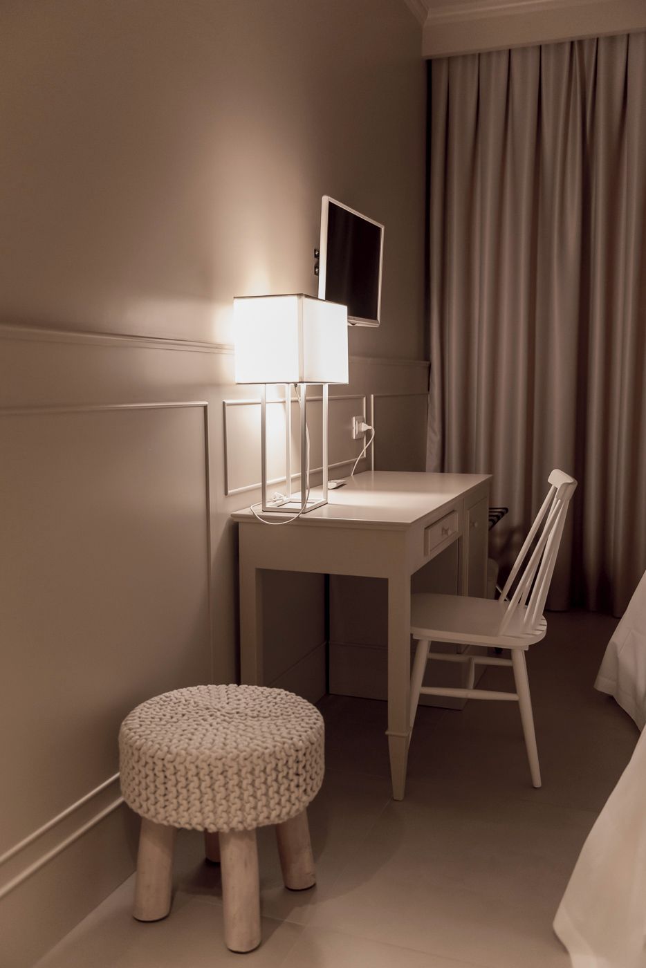 A hotel room with a white desk, a chair, a bedside table, and a lamp.