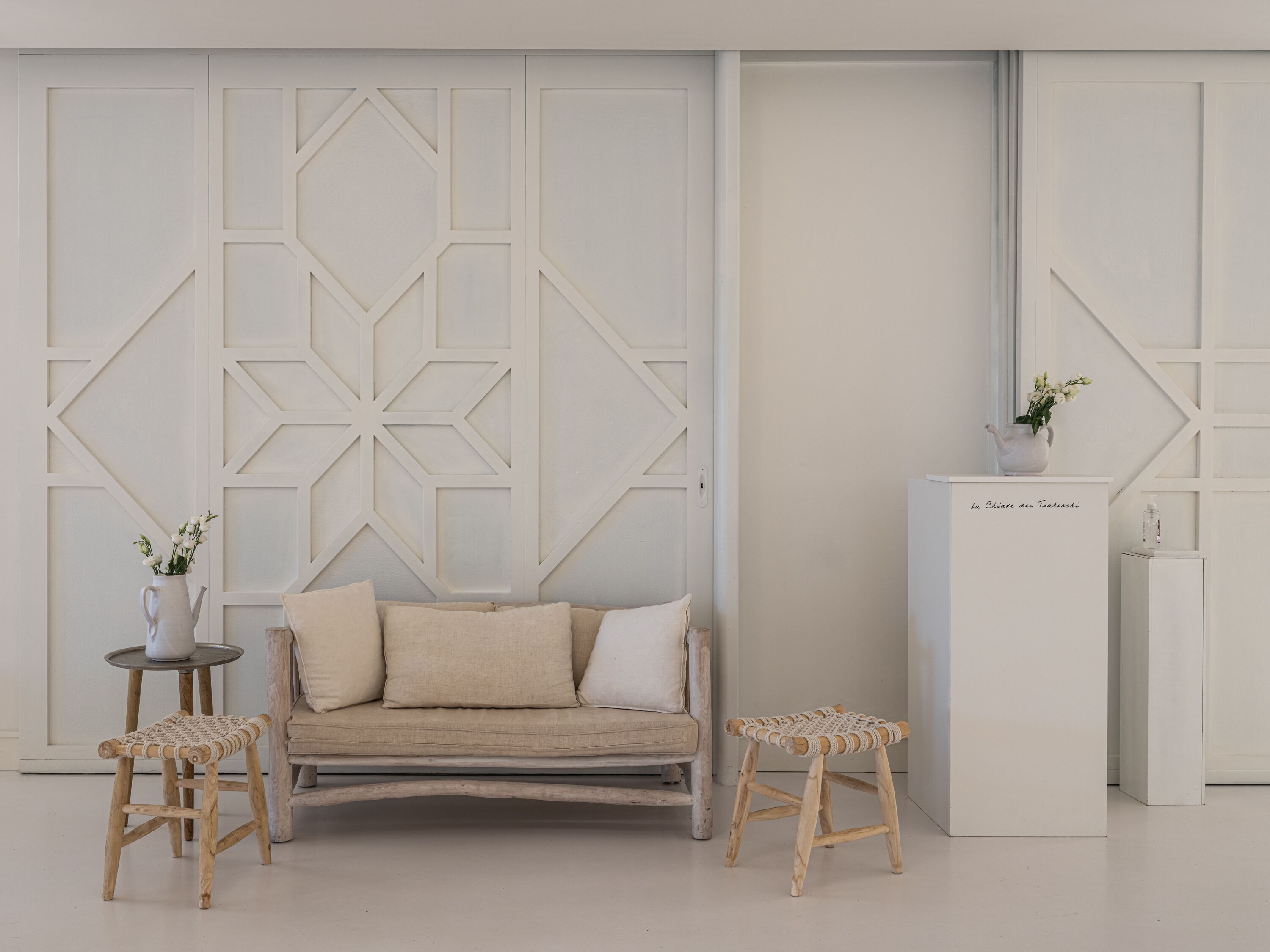 Una stanza dal design minimalista con un divano, due sgabelli in legno, un tavolino con un vaso e un mobile con una pianta.