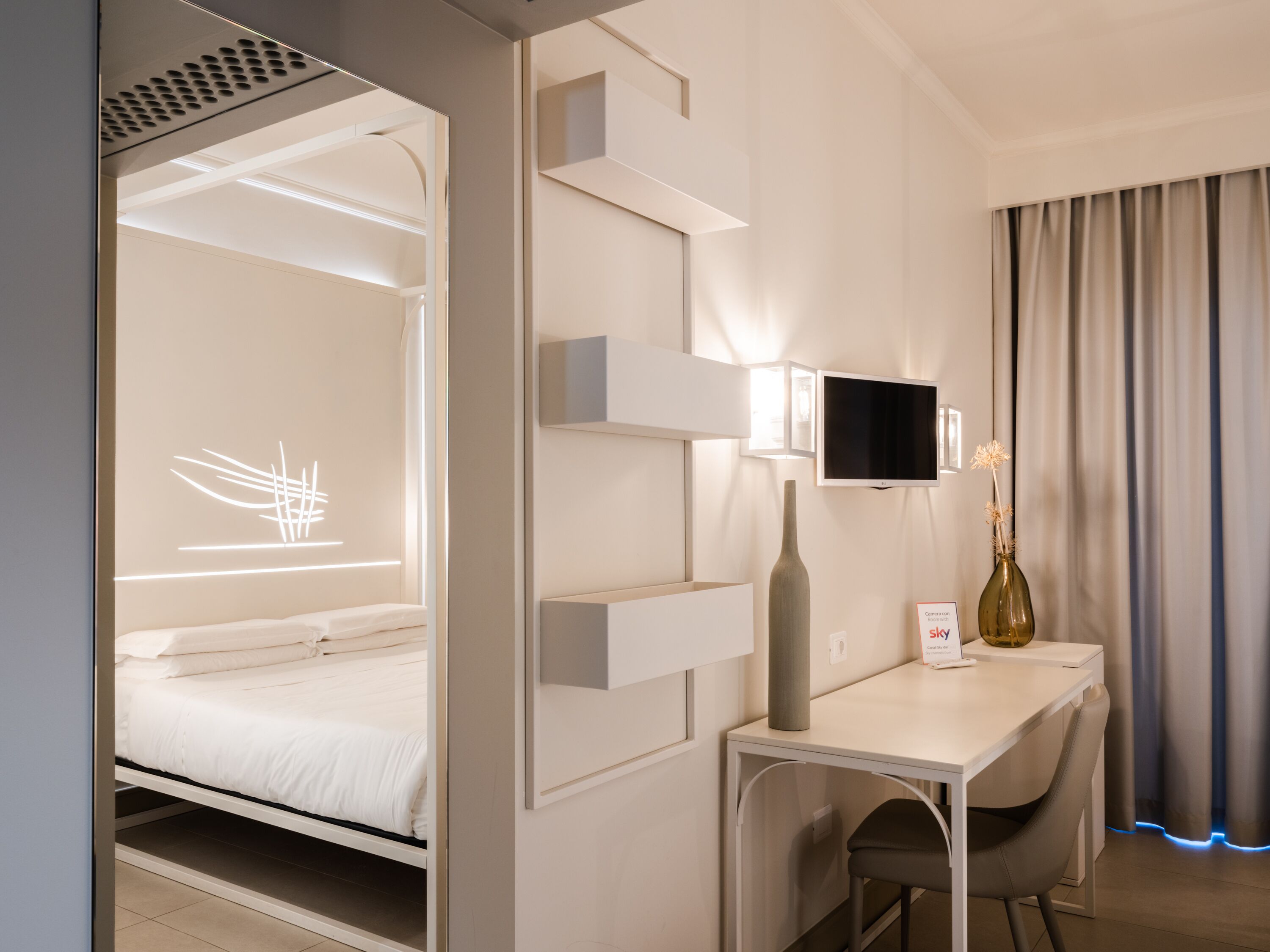 A modern hotel room with a bed, a desk with a chair, a television, and a vase with dried flowers.