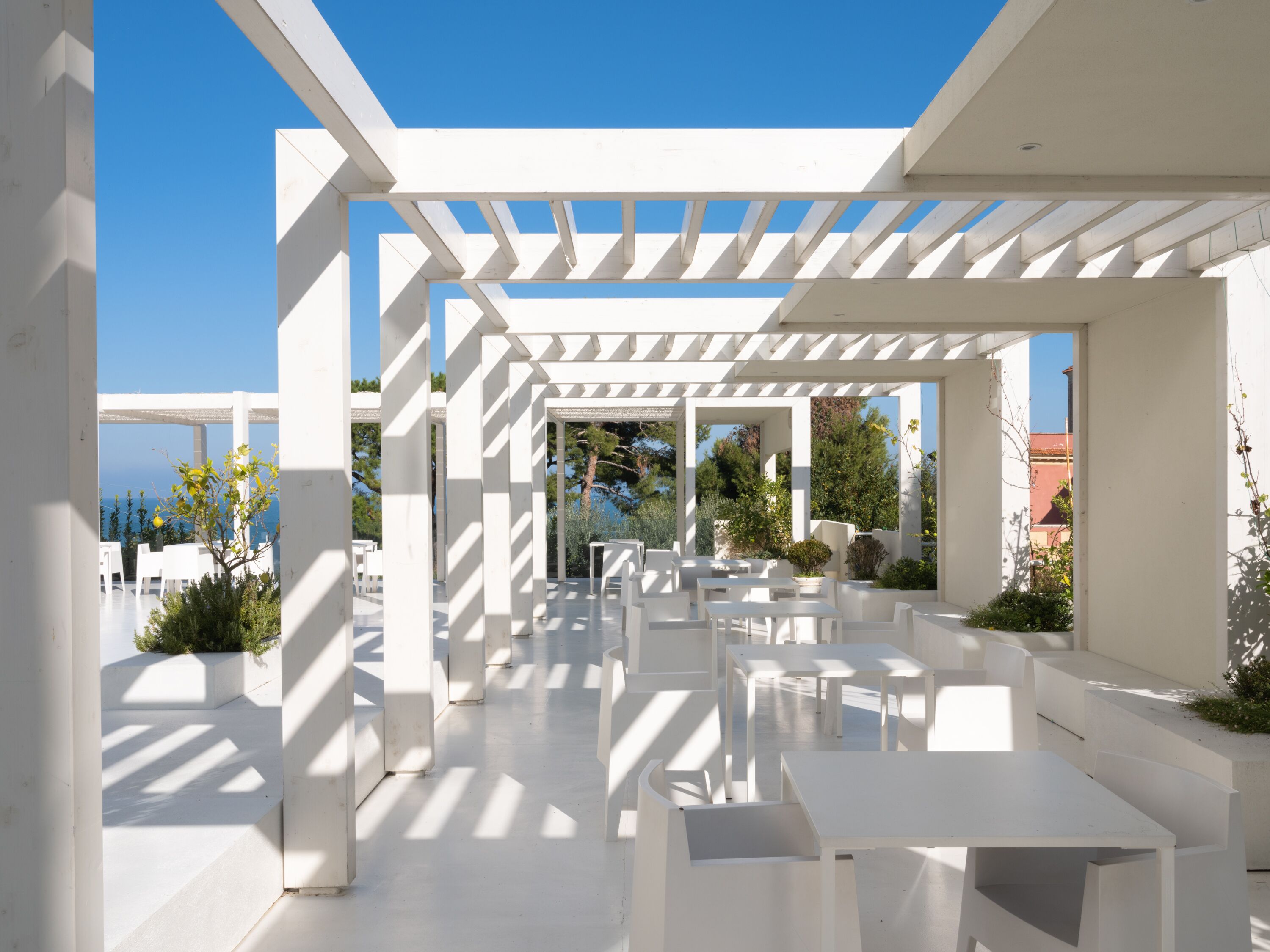 An outdoor patio area with white tables and chairs, a pergola structure, and a view of the sea.