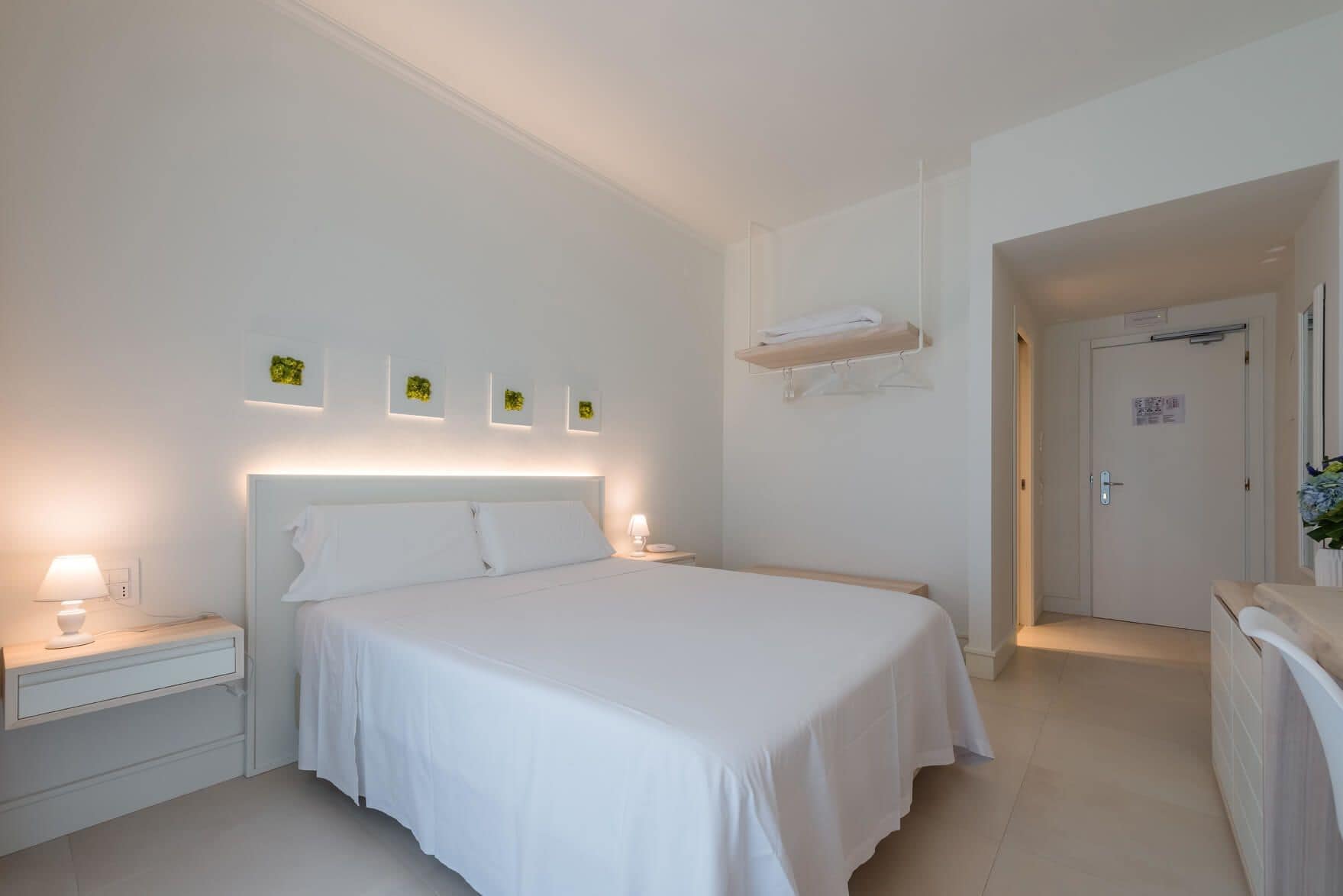 A modern hotel room with a large bed, bedside tables, a wall-mounted shelf, and a door leading to another room.