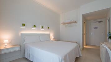 A modern hotel room with a large bed, bedside tables, a wall-mounted shelf, and a door leading to another room.