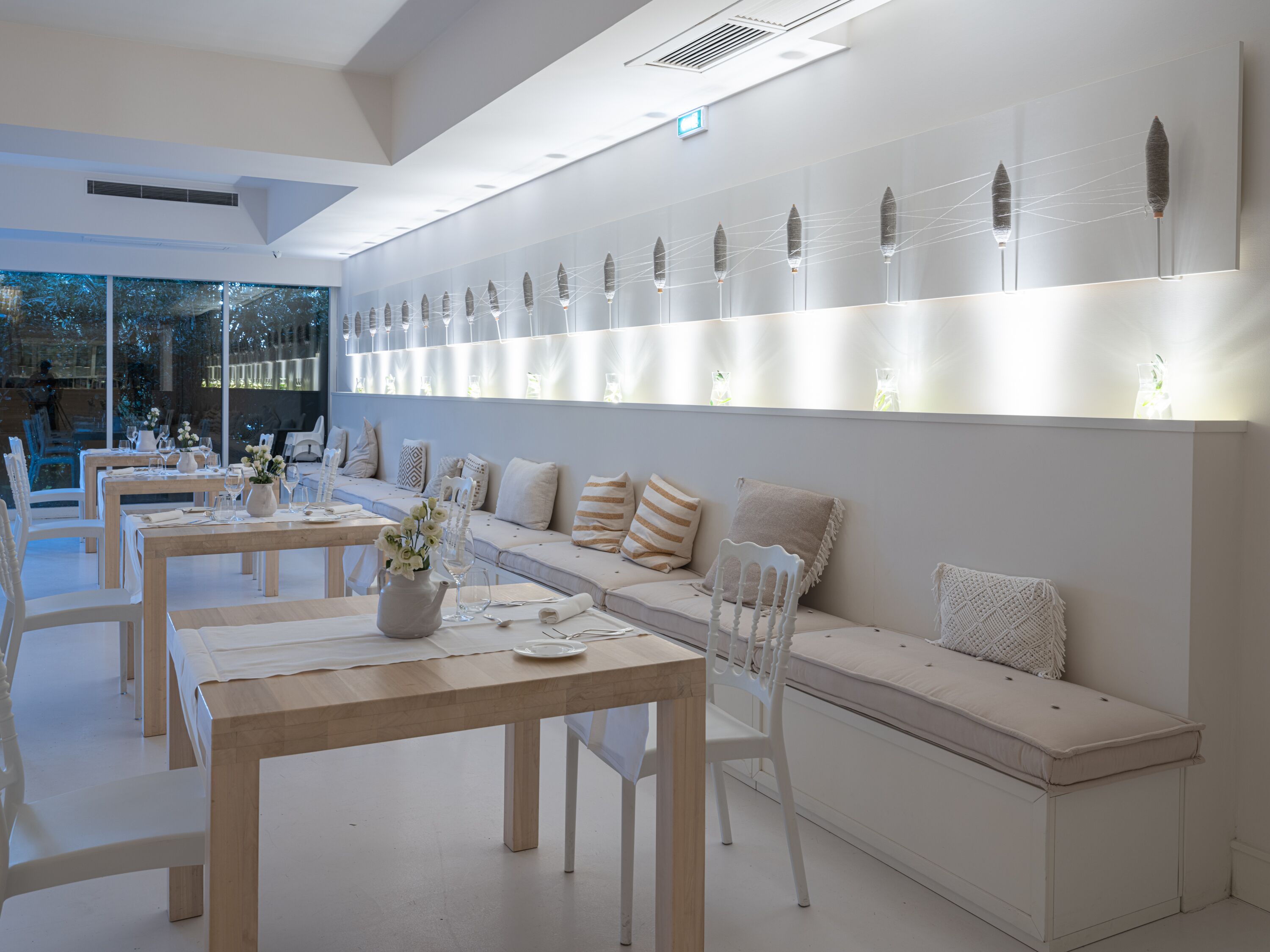 Un comedor moderno con mesas de madera y sillas blancas, un largo banco con cojines y una pared con detalles decorativos.