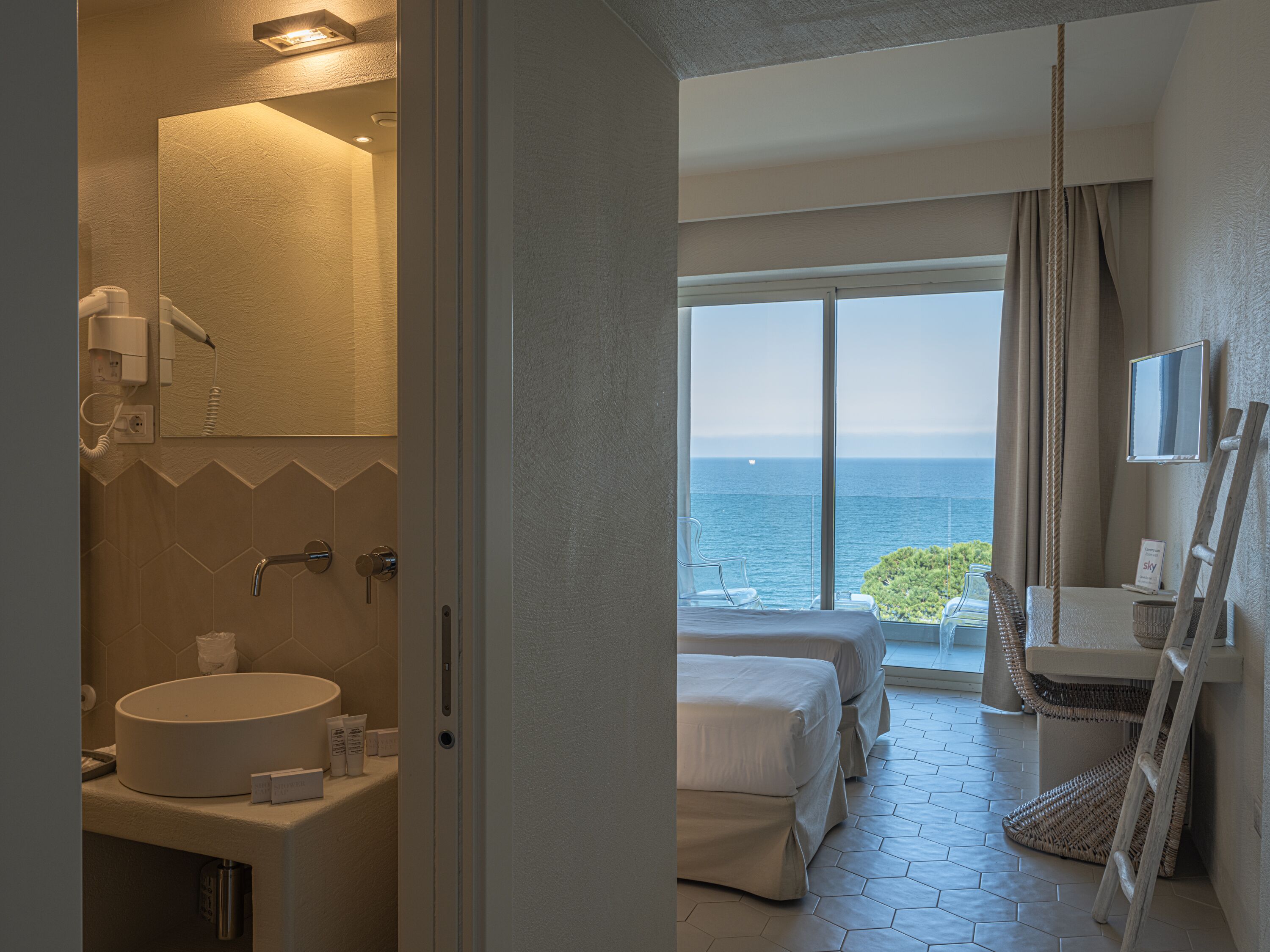 A hotel room with a large window overlooking the sea, a bathroom with a round sink, and a bed with white bedding.