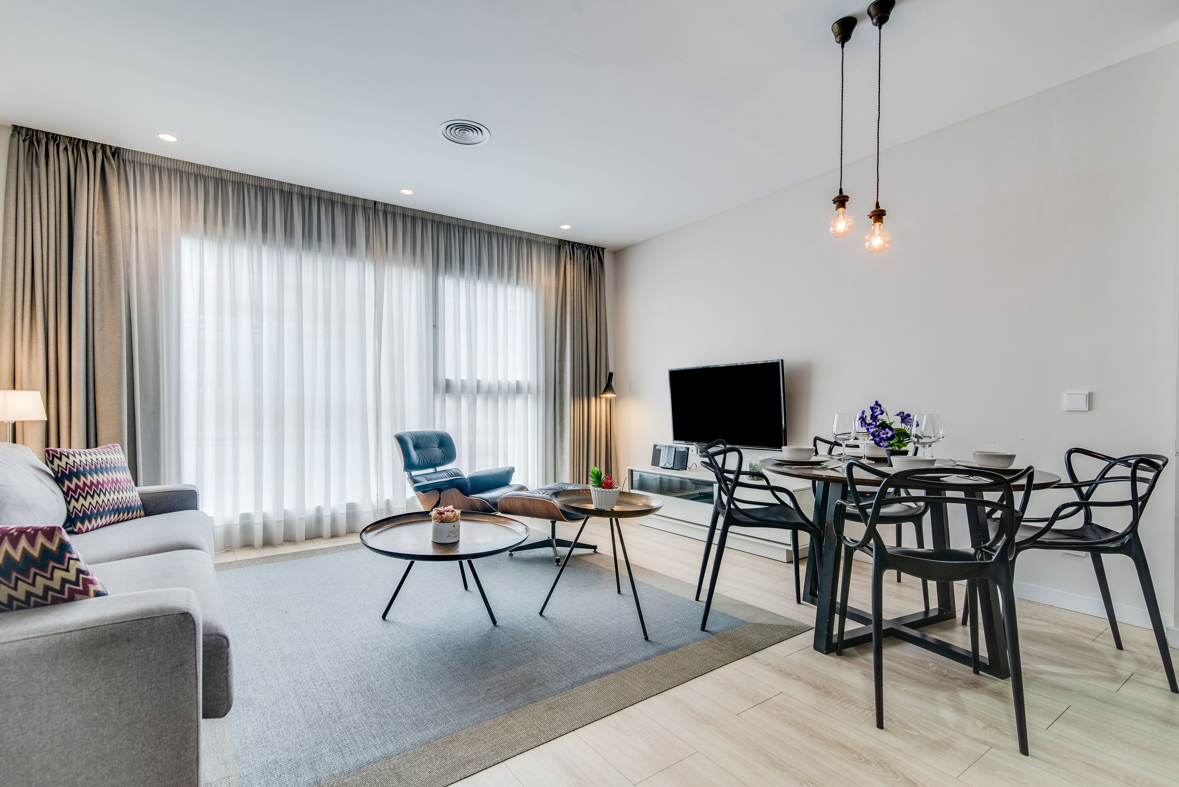 A modern living room with a sofa, coffee table, dining table, and chairs. A flat-screen TV is mounted on the wall.