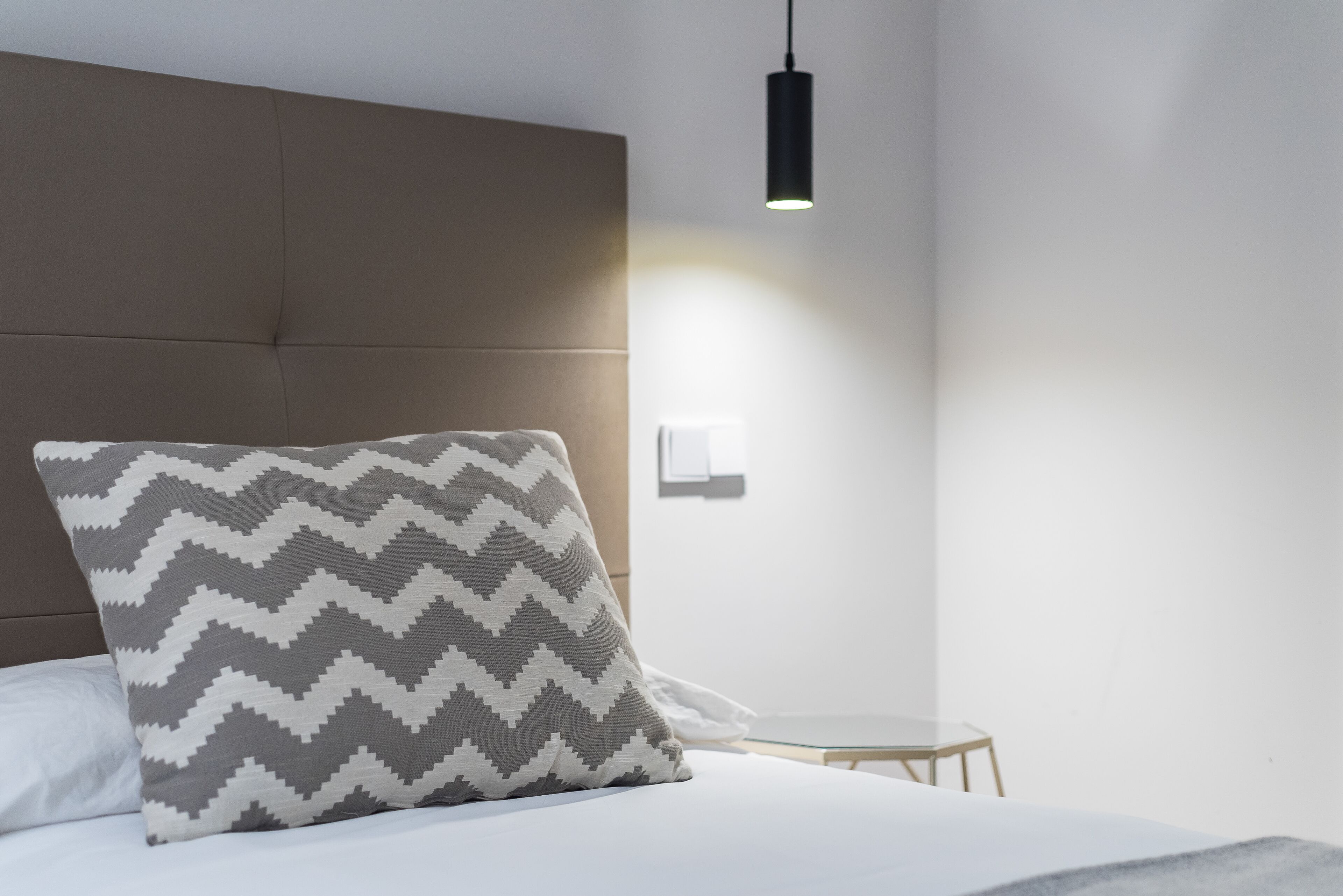 A neatly made bed with a patterned pillow, a bedside table, and a wall-mounted light fixture.