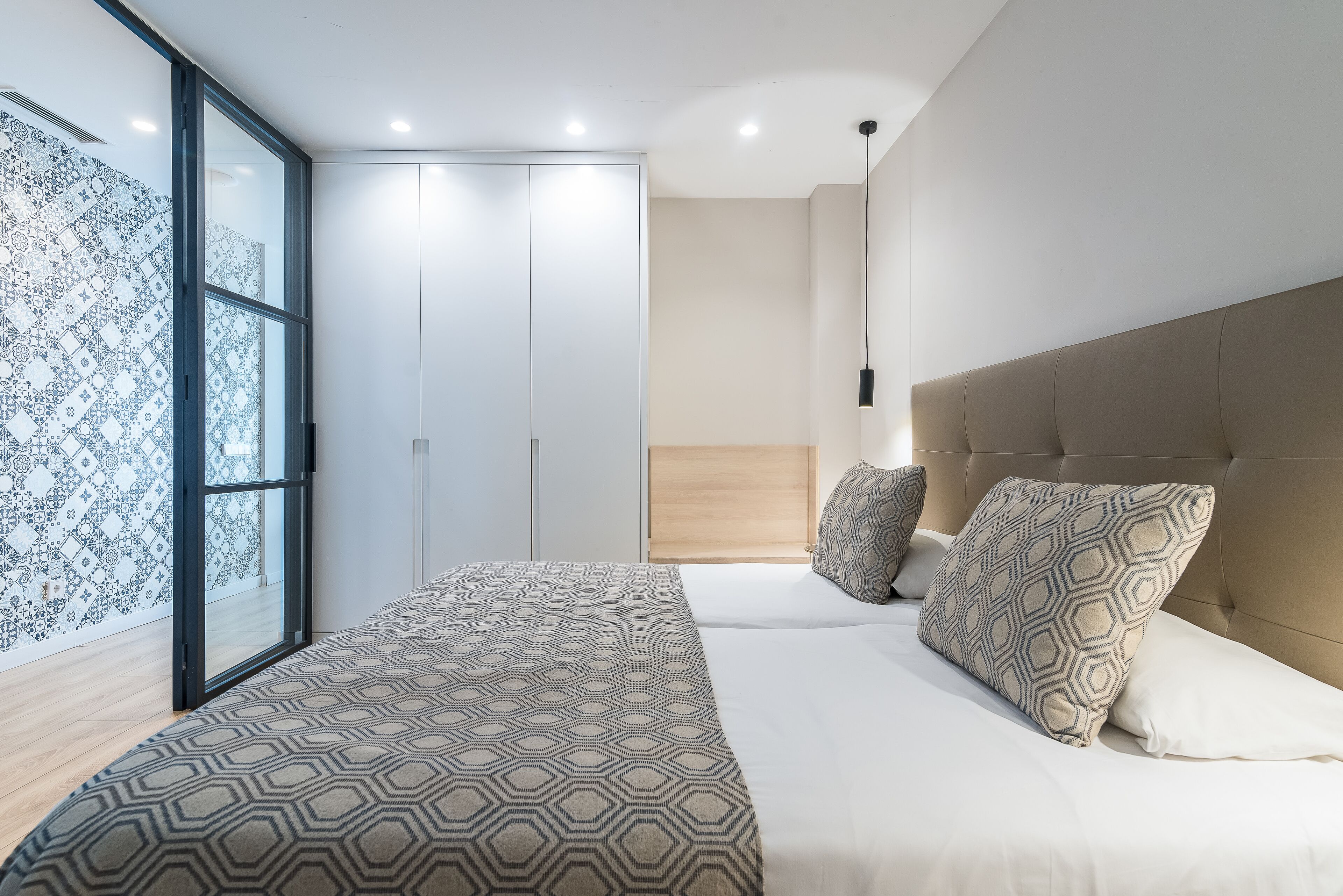 Ein modernes Schlafzimmer mit einem großen Bett, gemusterten Betttextilien, einem Kopfteil und einem großen Fenster mit Vorhängen.