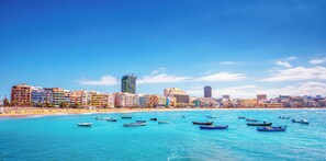 Un paisaje urbano costero con playa, numerosos barcos y varios edificios, incluyendo hoteles.