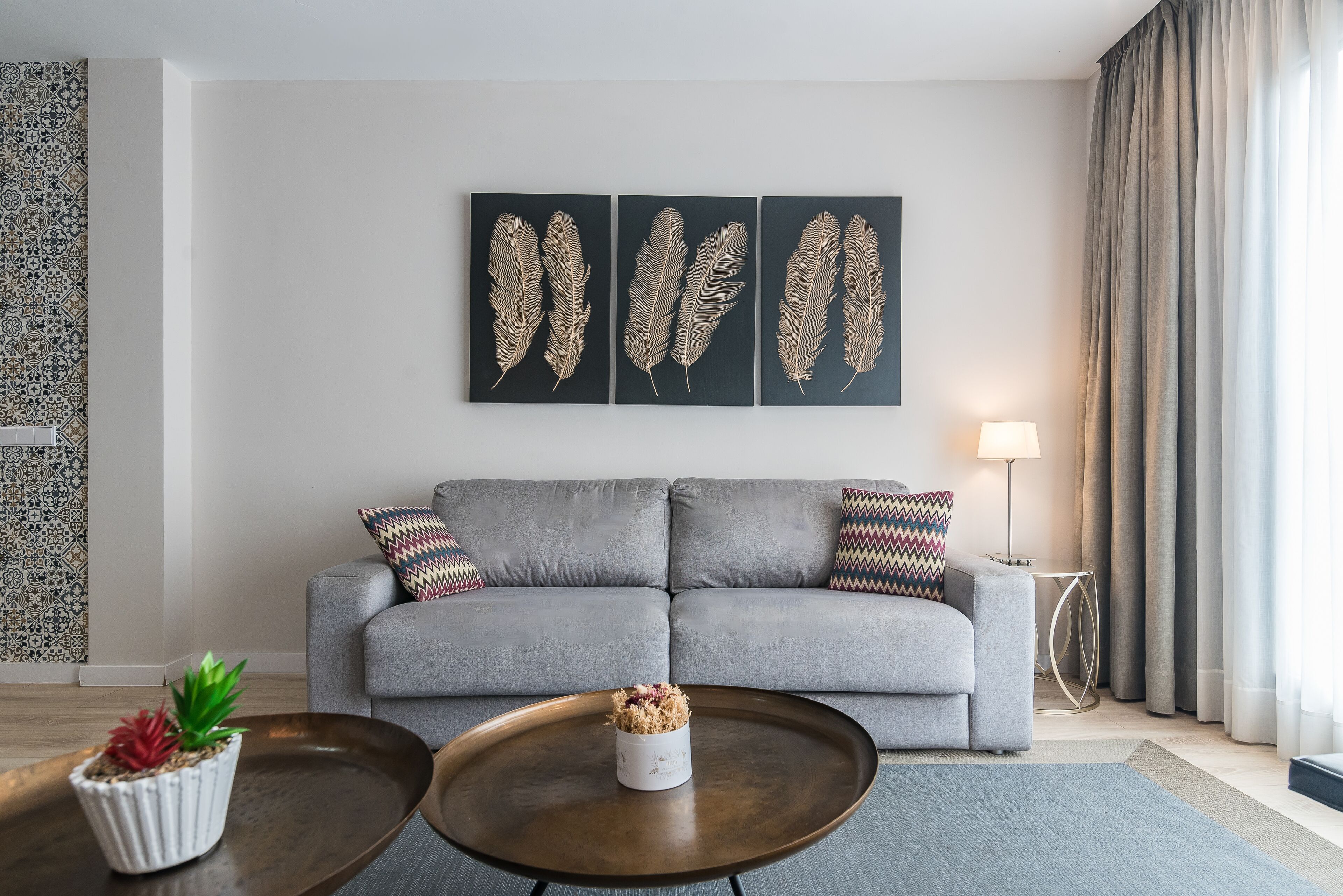 Ein modernes Wohnzimmer mit grauem Sofa, dekorativer Federwandgestaltung, einer kleinen Topfpflanze und einem runden Couchtisch.