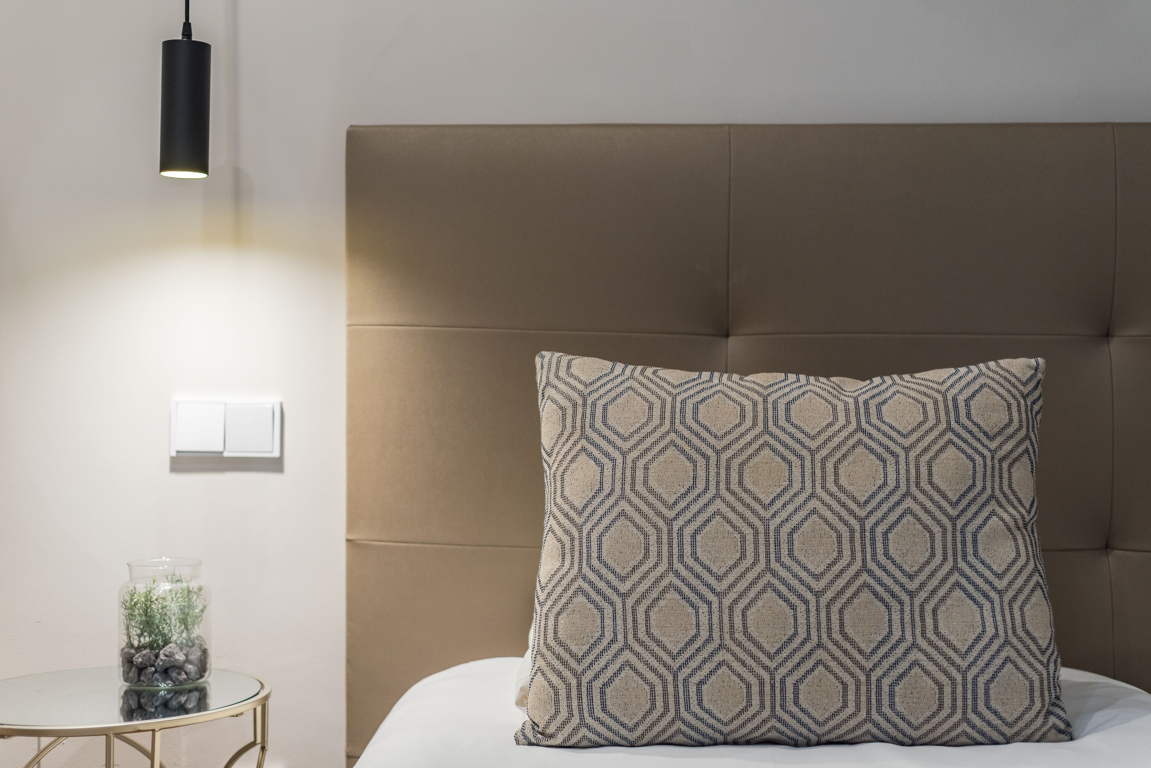 Una camera da letto con un letto, un comodino con un vasetto di vetro, un cuscino decorato e una lampada a sospensione.