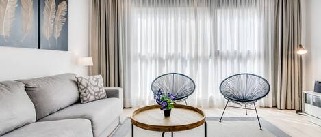 Una sala moderna con un sofá gris, dos sillas de estructura metálica negra, una mesa de centro de madera con un jarrón de flores y un ventanal con cortinas translúcidas.