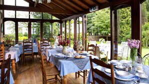 Un área de comedor al aire libre con mesas y sillas de madera, cubierta por un techo de madera y rodeada de abundante vegetación.