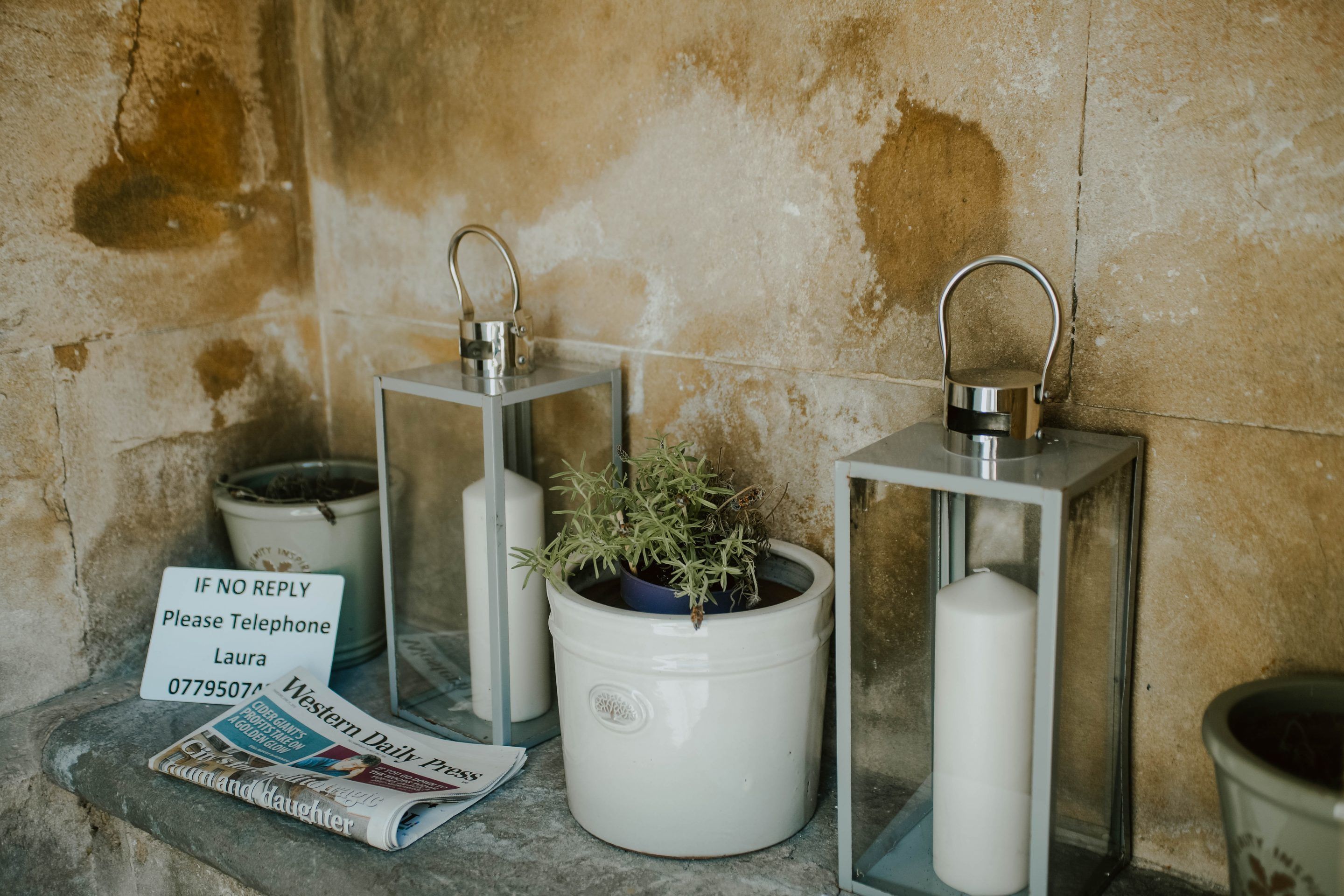 Two white lanterns with metal frames and a potted plant in a white bucket, a sign with a phone number, and a newspaper on a stone ledge against a textured wall.