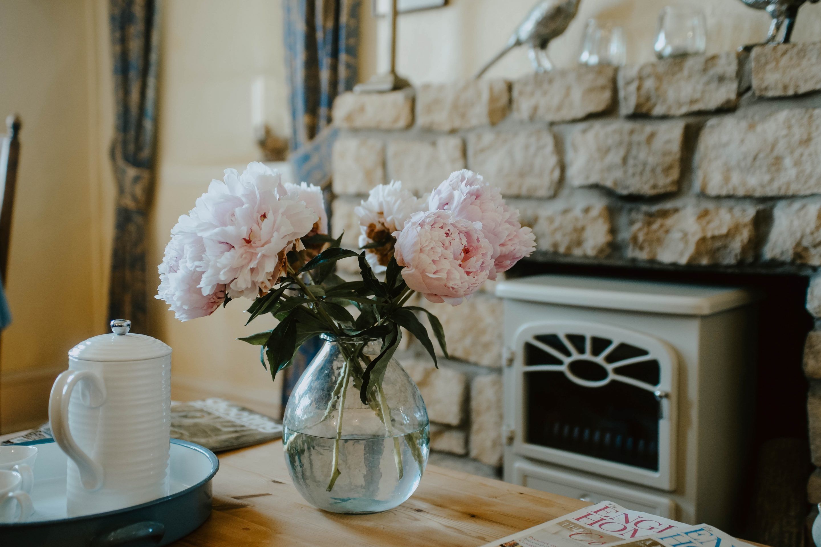 On voit clairement une pièce avec un mur en pierre, une cheminée blanche et un vase rempli de fleurs posé sur une table en bois.