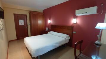 Ein Hotelzimmer mit einem Einzelbett, einem Holzschrank, einem Schreibtisch mit Lampe und einer Klimaanlage an der Wand.