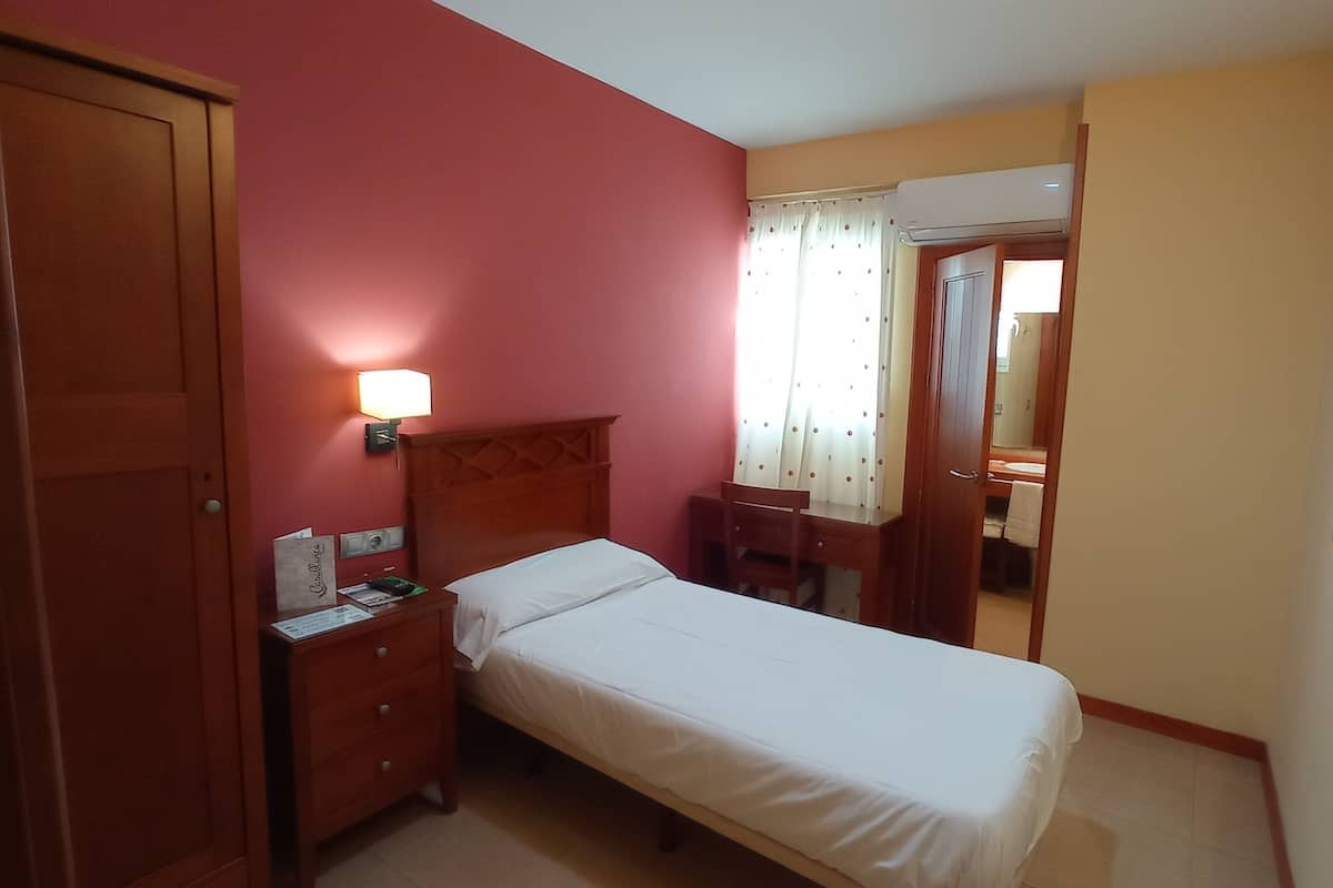 Ein Hotelzimmer mit einem Einzelbett, einem Nachttisch aus Holz mit Lampe, einem Schreibtisch mit Stuhl und einem Spiegel.