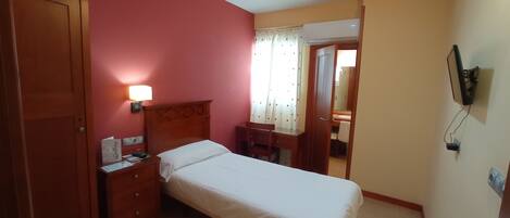 Habitación de hotel con una cama individual, una mesita de noche de madera con lámpara, un escritorio con silla y un espejo.