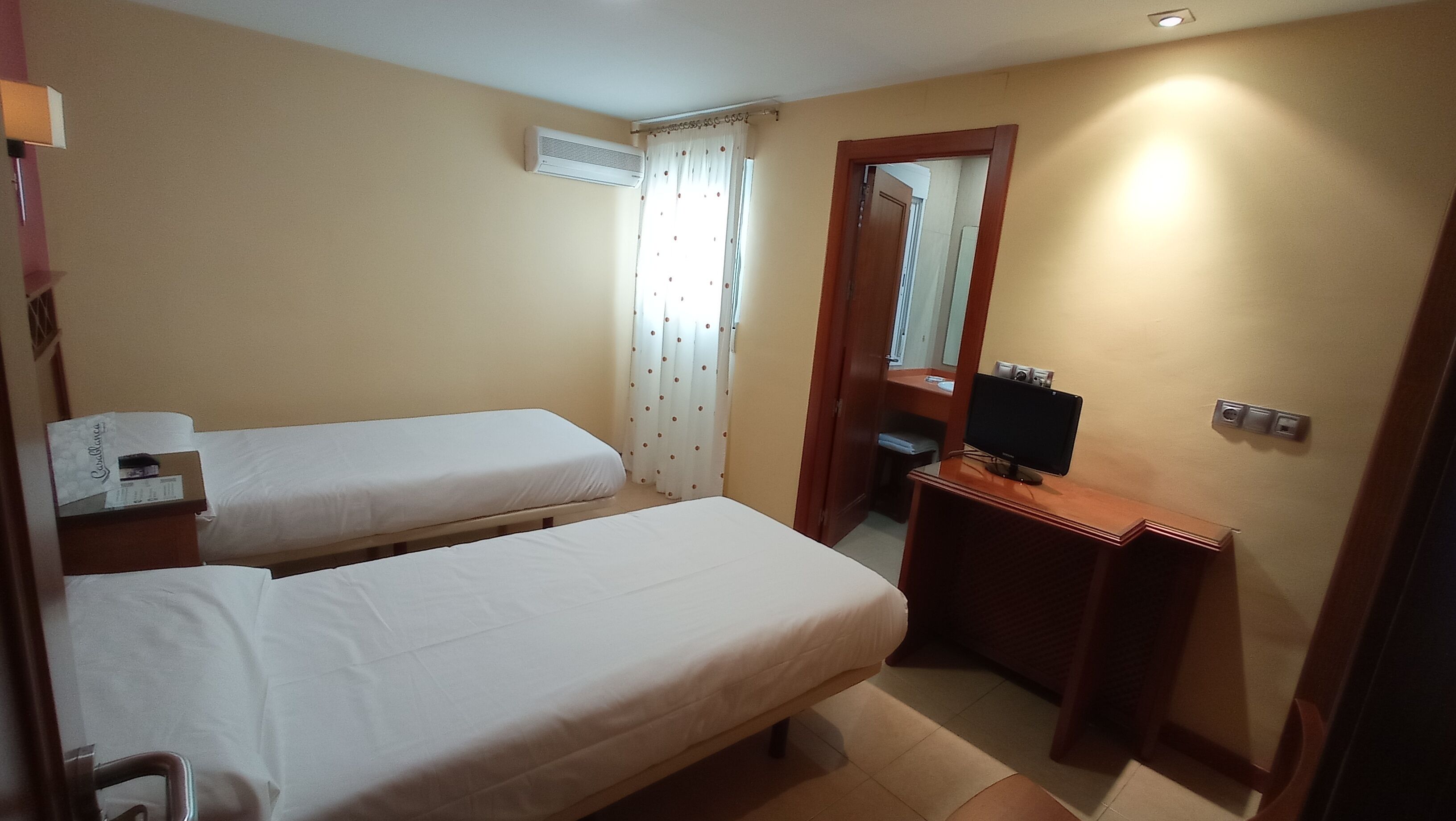 A small hotel room with two single beds, a desk with a computer, a mirror, and a window with curtains.