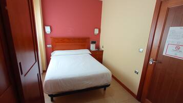 A hotel room with a single bed, a nightstand, a red wall, and a door with a no-smoking sign.