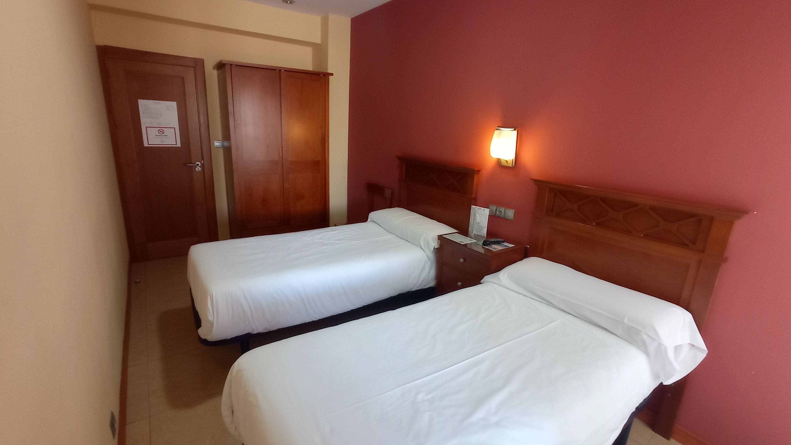 Habitación de hotel con dos camas individuales, un armario de madera, una mesita de noche con lámpara y una pared roja.