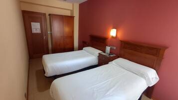 A hotel room with two single beds, a wooden wardrobe, a nightstand with a lamp, and a red wall.