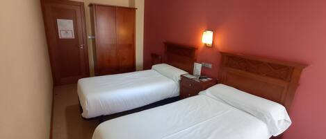 Habitación de hotel con dos camas individuales, un armario de madera, una mesita de noche con lámpara y una pared roja.