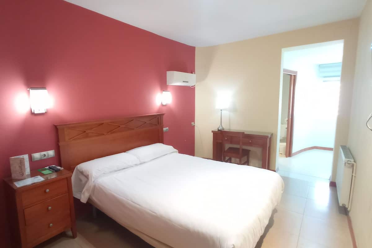 Ein Hotelzimmer mit einem Bett, einem Schreibtisch und einem Stuhl. Ein Nachttisch mit Lampe und Steckdose ist vorhanden. Der Raum hat eine rote Wand und eine beigefarbene Wand.