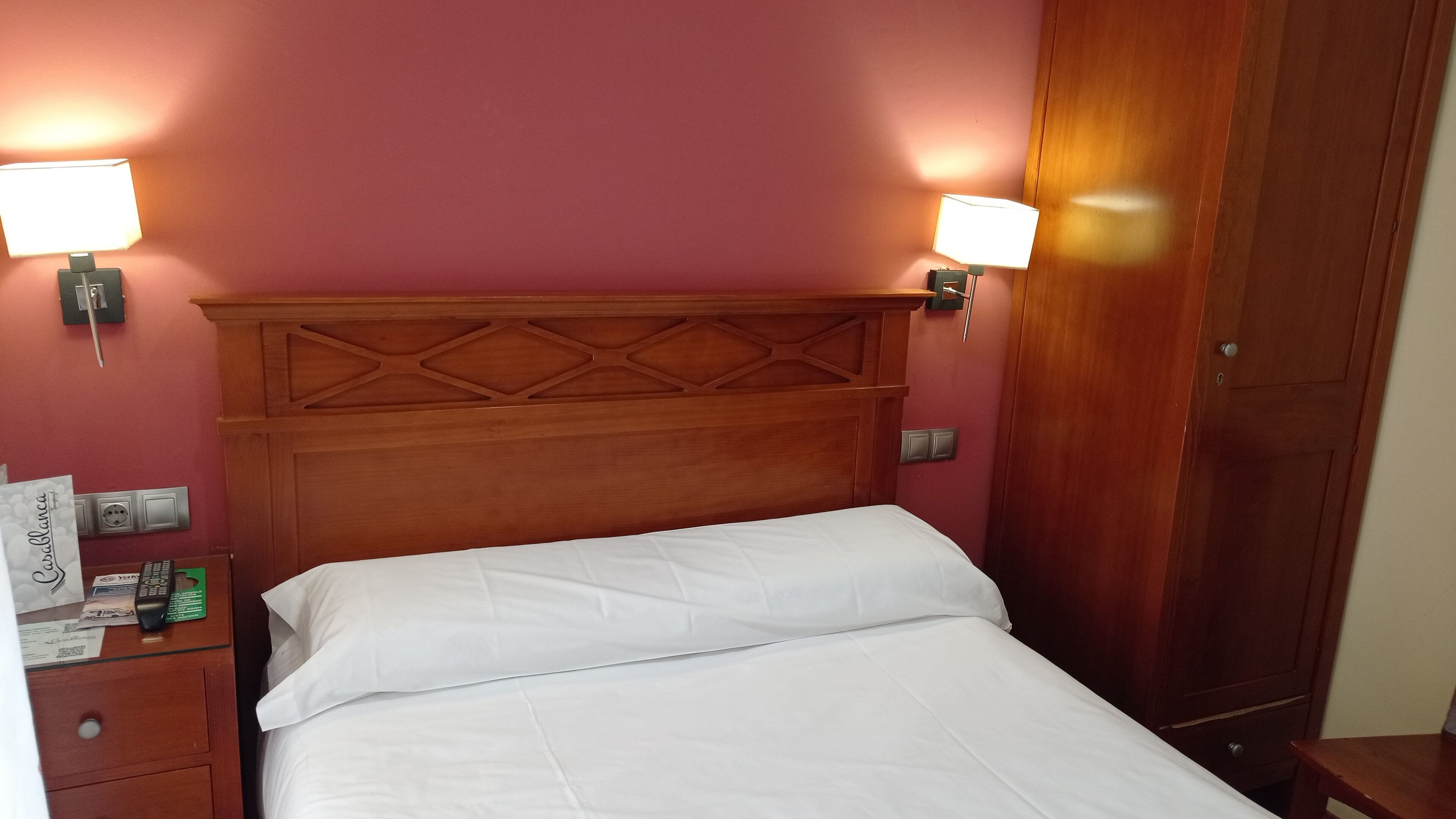 A hotel room with a wooden headboard, two bedside lamps, a bed with white linens, a wooden nightstand, and a wooden wardrobe.