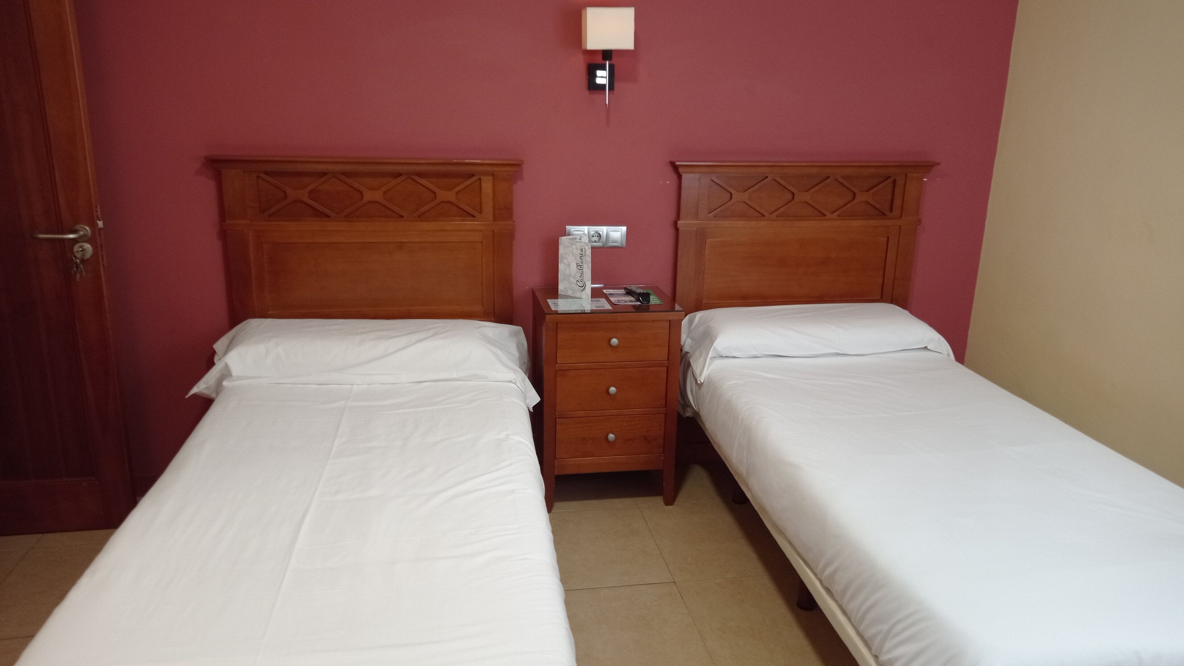 Una habitació amb dos llits individuals, una tauleta de nit de fusta amb una làmpada i un termòstat fixat a la paret.