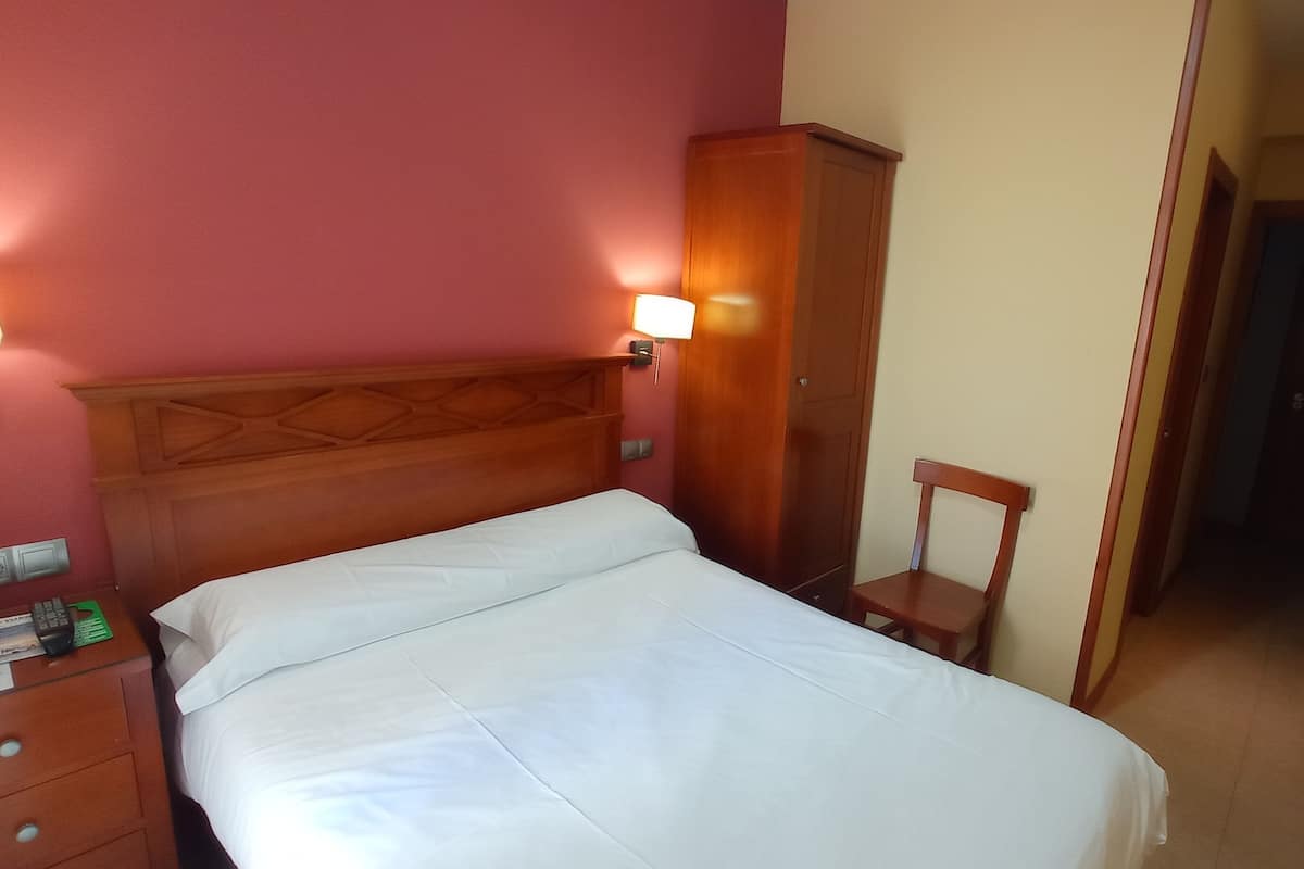 Ein Hotelzimmer mit Holzkopfteil, einem Einzelbett, einem Nachttisch, einer Nachttischlampe, einem Kleiderschrank und einem Sessel.