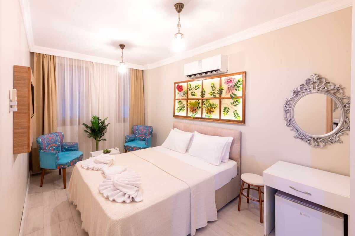 A hotel room with a bed, two chairs, a desk, a mirror, and a wall-mounted air conditioning unit.