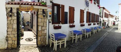 Un vicolo stretto con file di tavoli e sedie all'aperto, un arco in pietra che fa da ingresso e un edificio con persiane di legno e piante pensili.