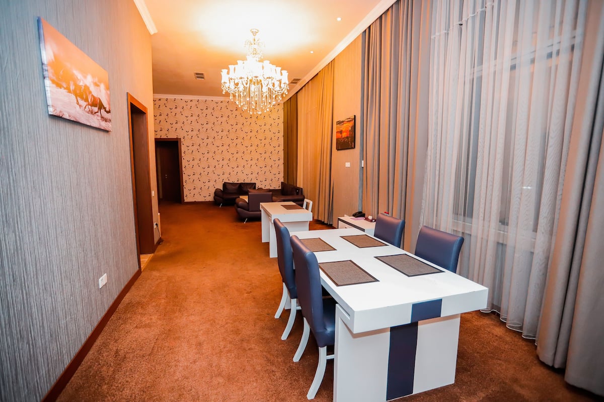 A hotel dining area with a long table, chairs, a chandelier, and a view through sheer curtains.