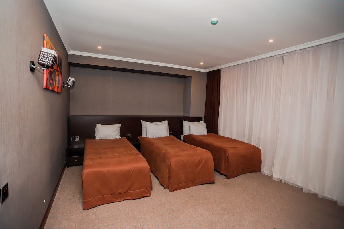 A hotel room with three single beds, a large window with white curtains, a wall-mounted artwork, and a ceiling light.