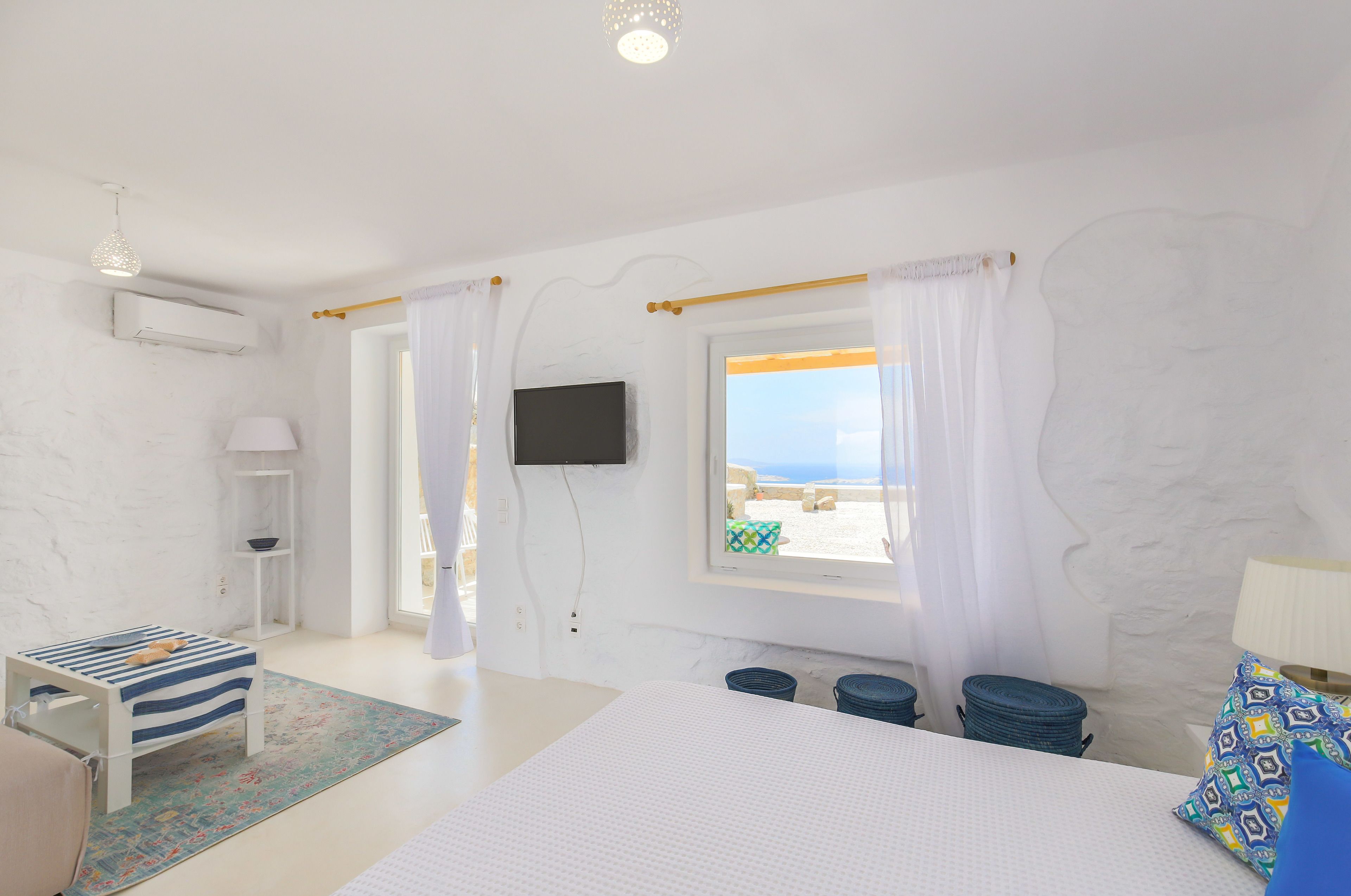 Ein Schlafzimmer mit einem Bett, einem an der Wand montierten Fernseher, einem Fenster mit Vorhängen, einem kleinen Tisch mit gestreifter Tischdecke und einem farbenfrohen Teppich.