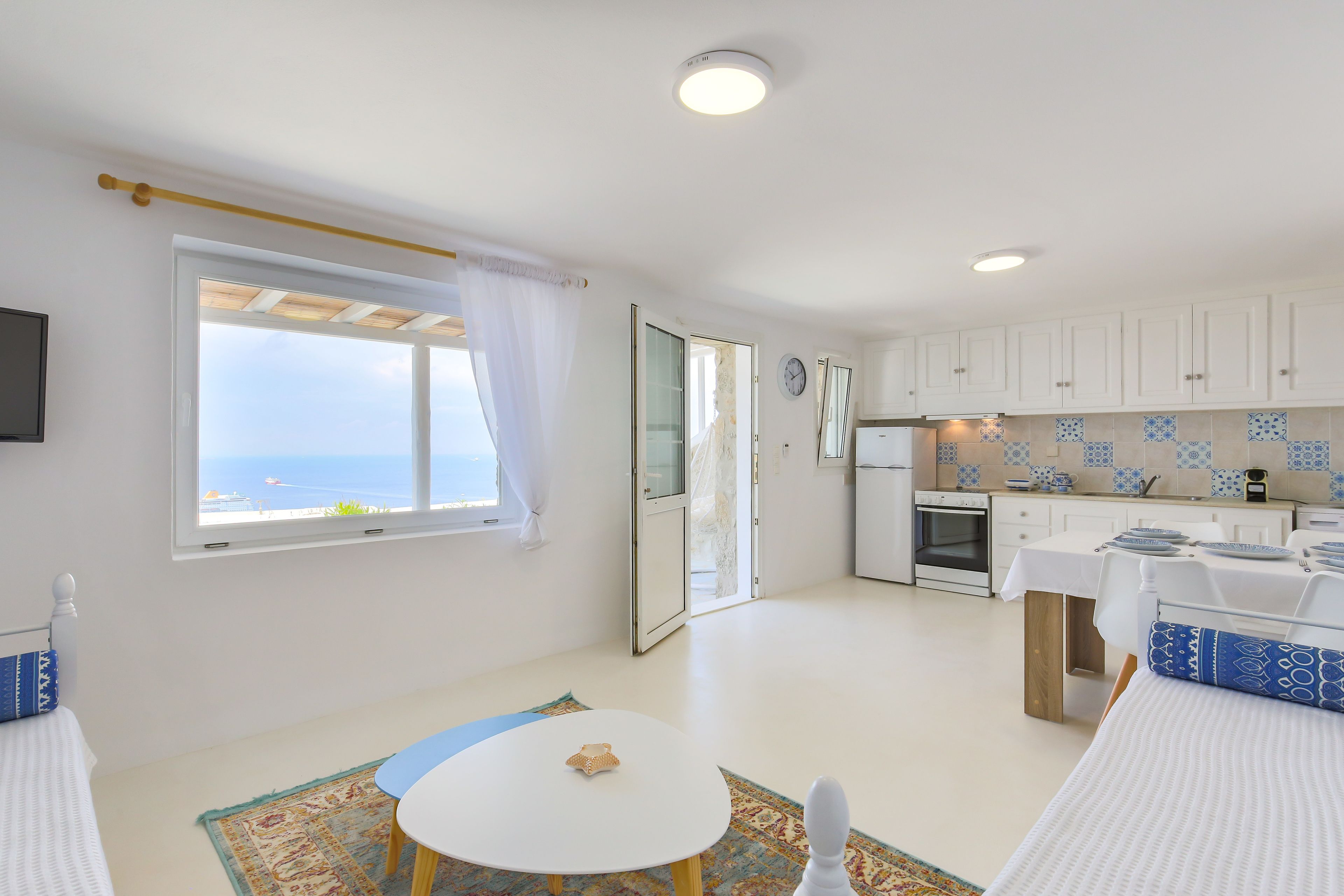 A modern, well-lit room with a large window, a small round table, a bed with a blue pillow, and a kitchen area with white cabinets and blue-tiled backsplash.