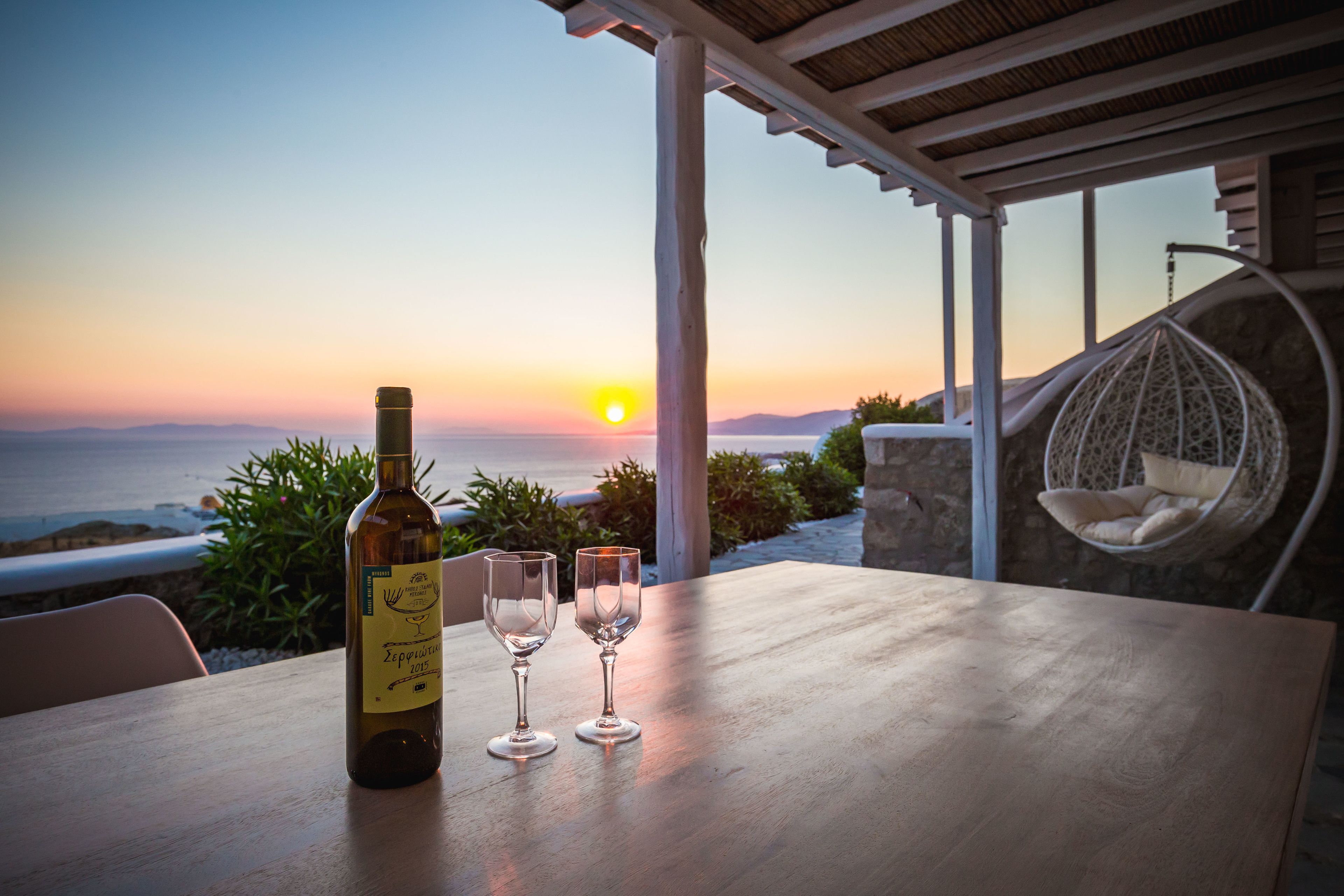 A table with a wine bottle and two glasses, overlooking a sunset and the sea.