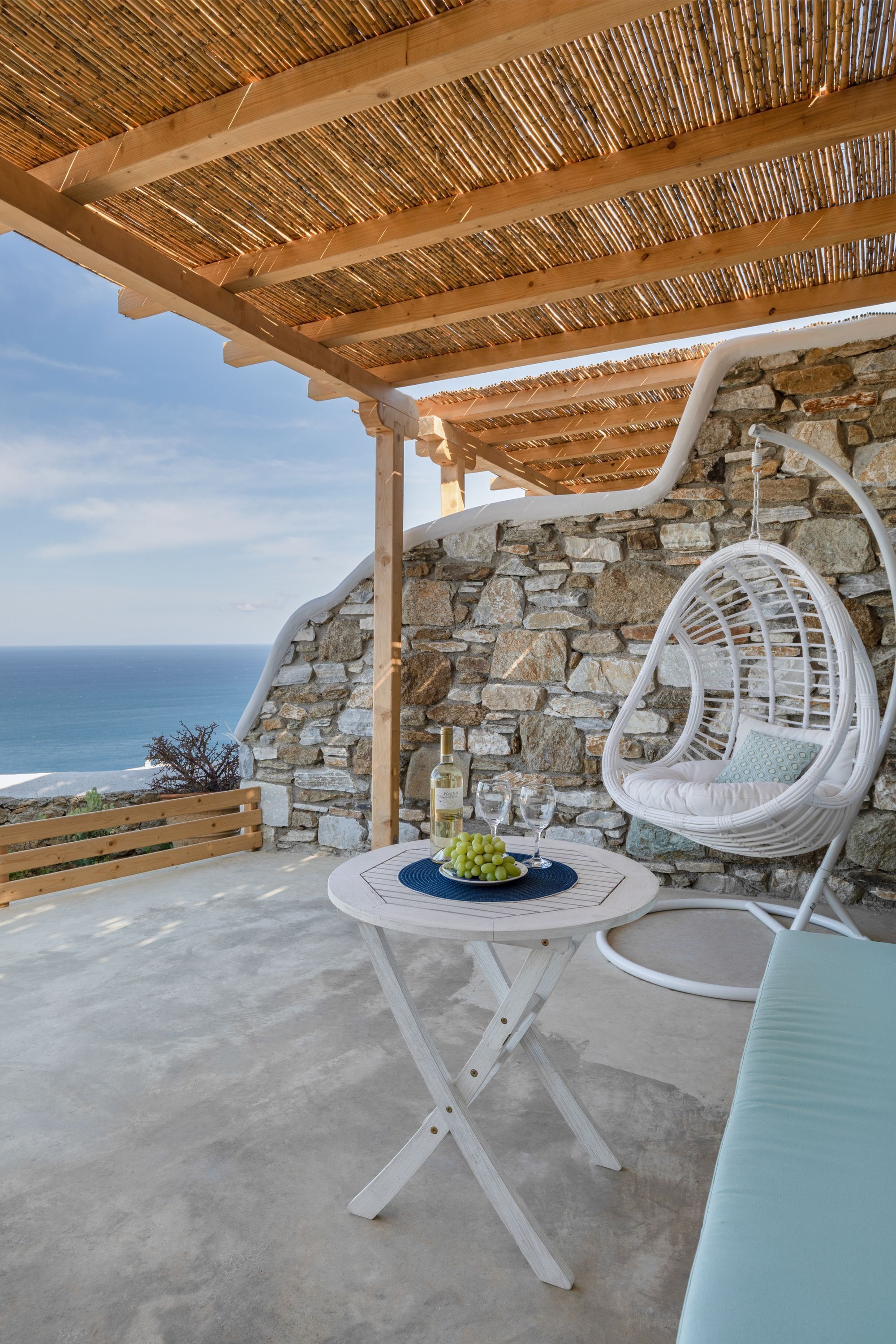 A patio with a stone wall, a hanging chair, a table with a bowl of fruit and glasses, and a view of the sea.
