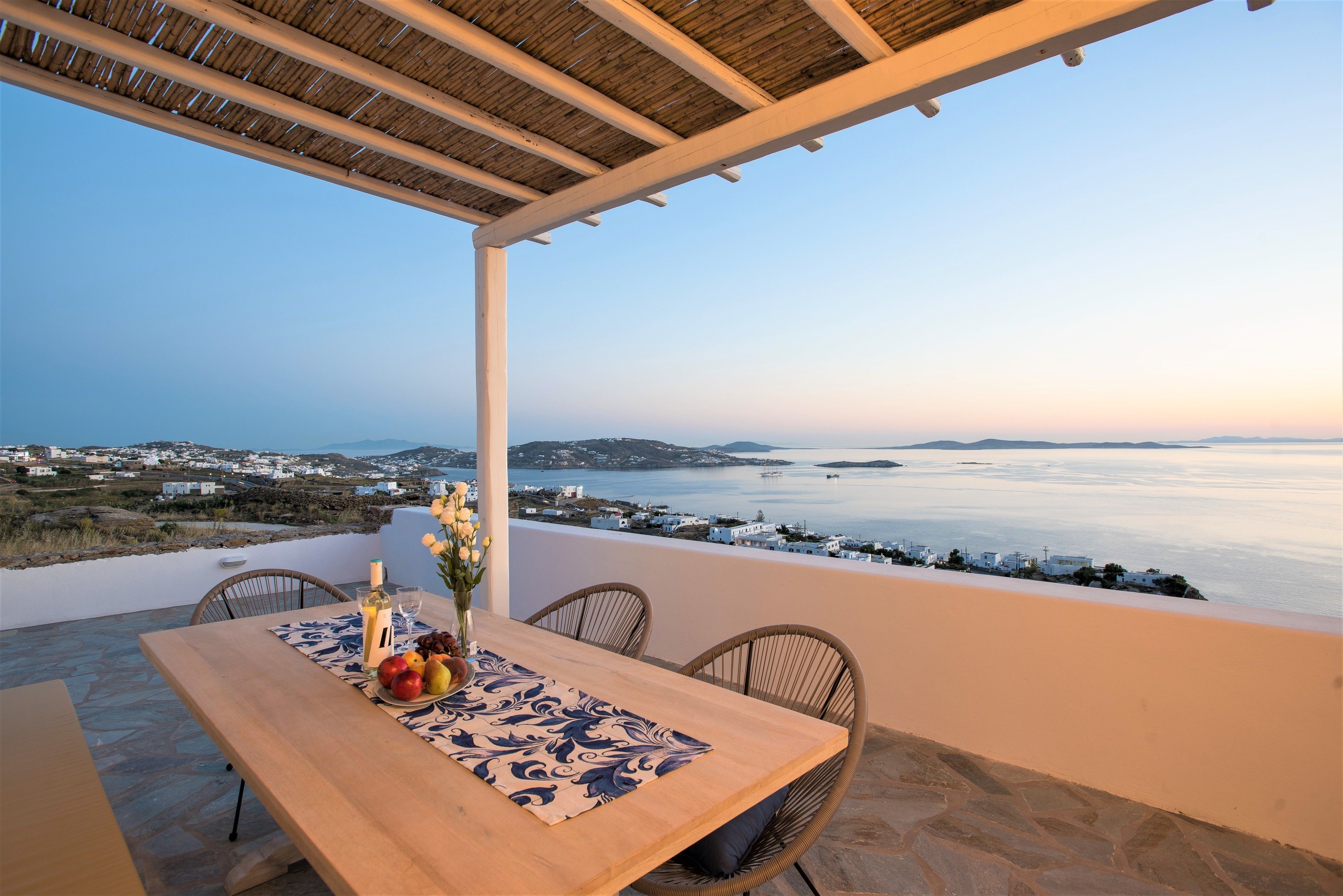 Una mesa dispuesta con frutas y una copa de vino, con vistas a la costa, edificios y el mar.