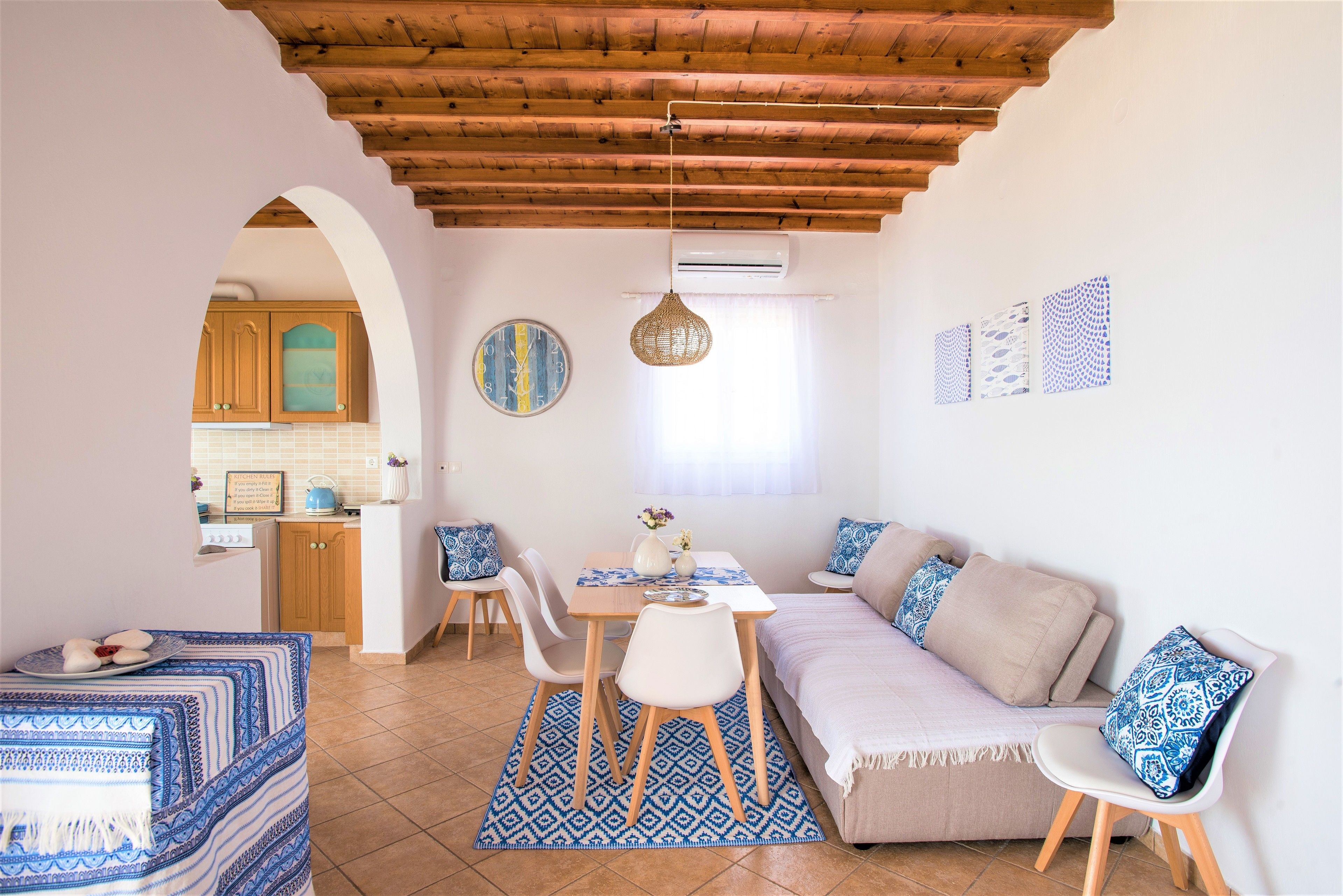 A cozy living area with a sofa, dining table, and chairs, an archway leading to a kitchen, and a ceiling with exposed wooden beams.
