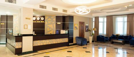 A hotel reception area with a reception desk, a clock, a seating area with sofas, and a large chandelier.