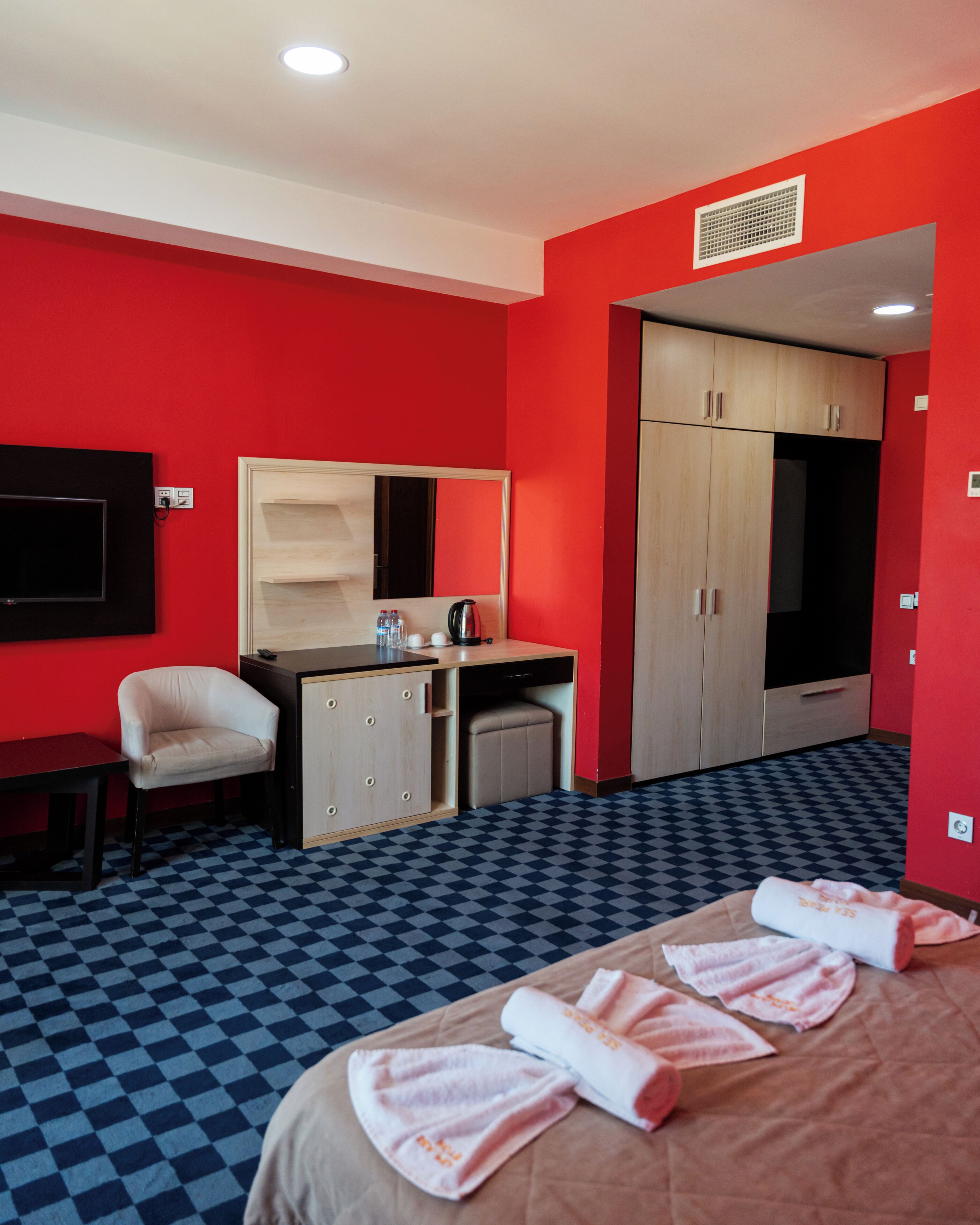 A hotel room with a checkered floor, a bed with towels, a desk with a mirror, a chair, and a wardrobe.