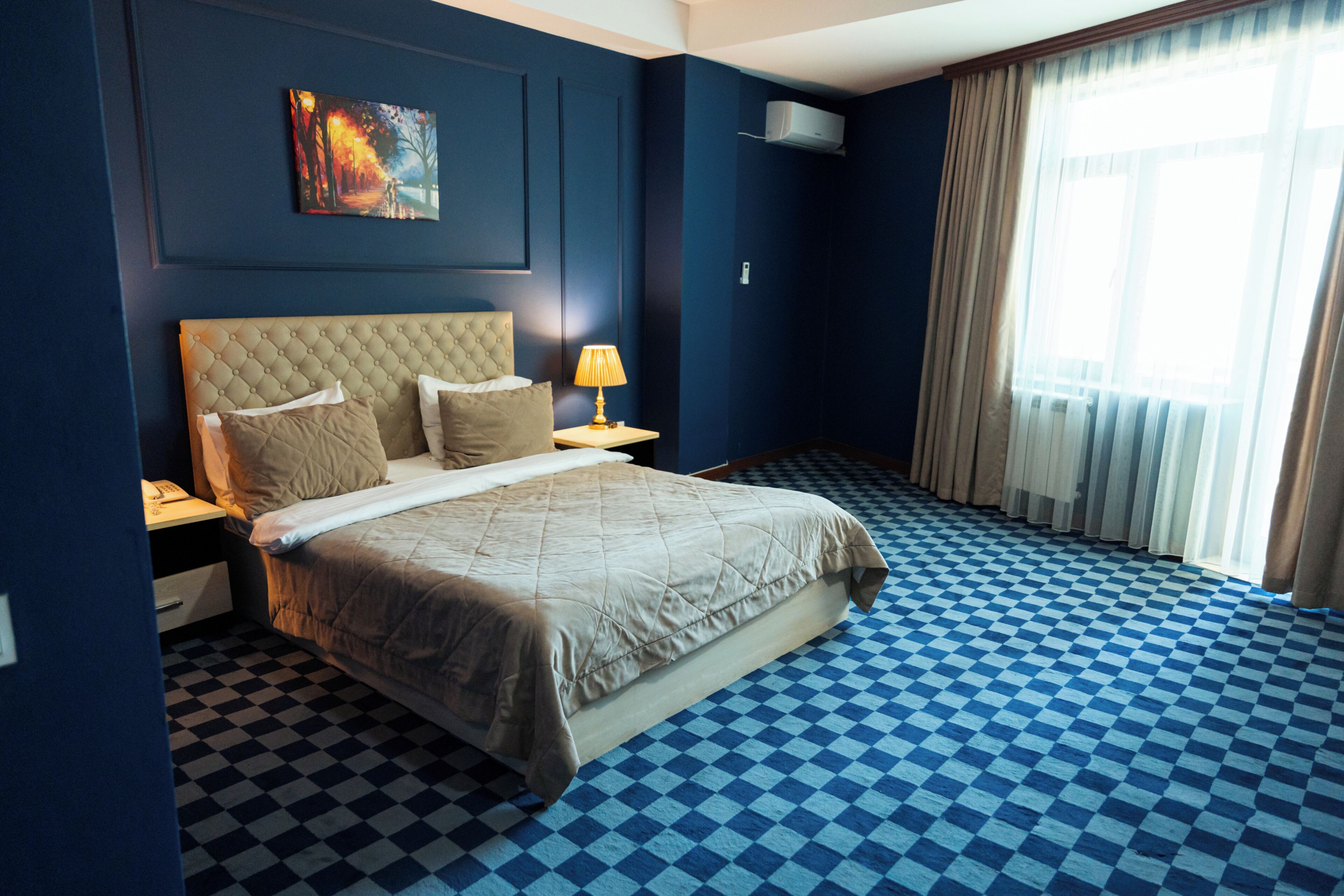 A hotel room with a checkered floor, a bed with a quilted headboard, a nightstand with a lamp, and a painting on the wall.