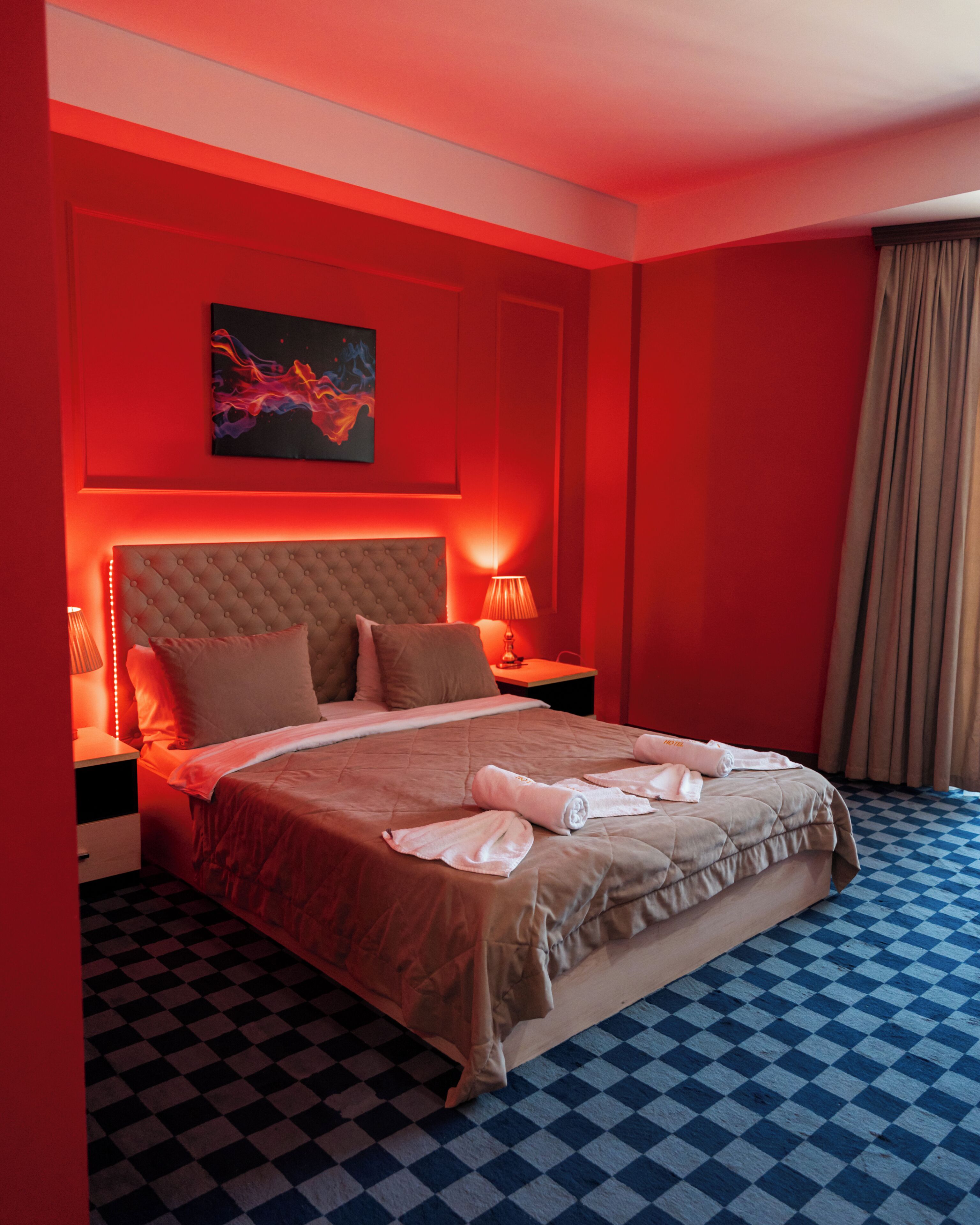 Une chambre d’hôtel avec un sol à carreaux, une tête de lit capitonnée, un lit avec des oreillers et une couverture, un tableau au mur et une table de chevet avec une lampe.