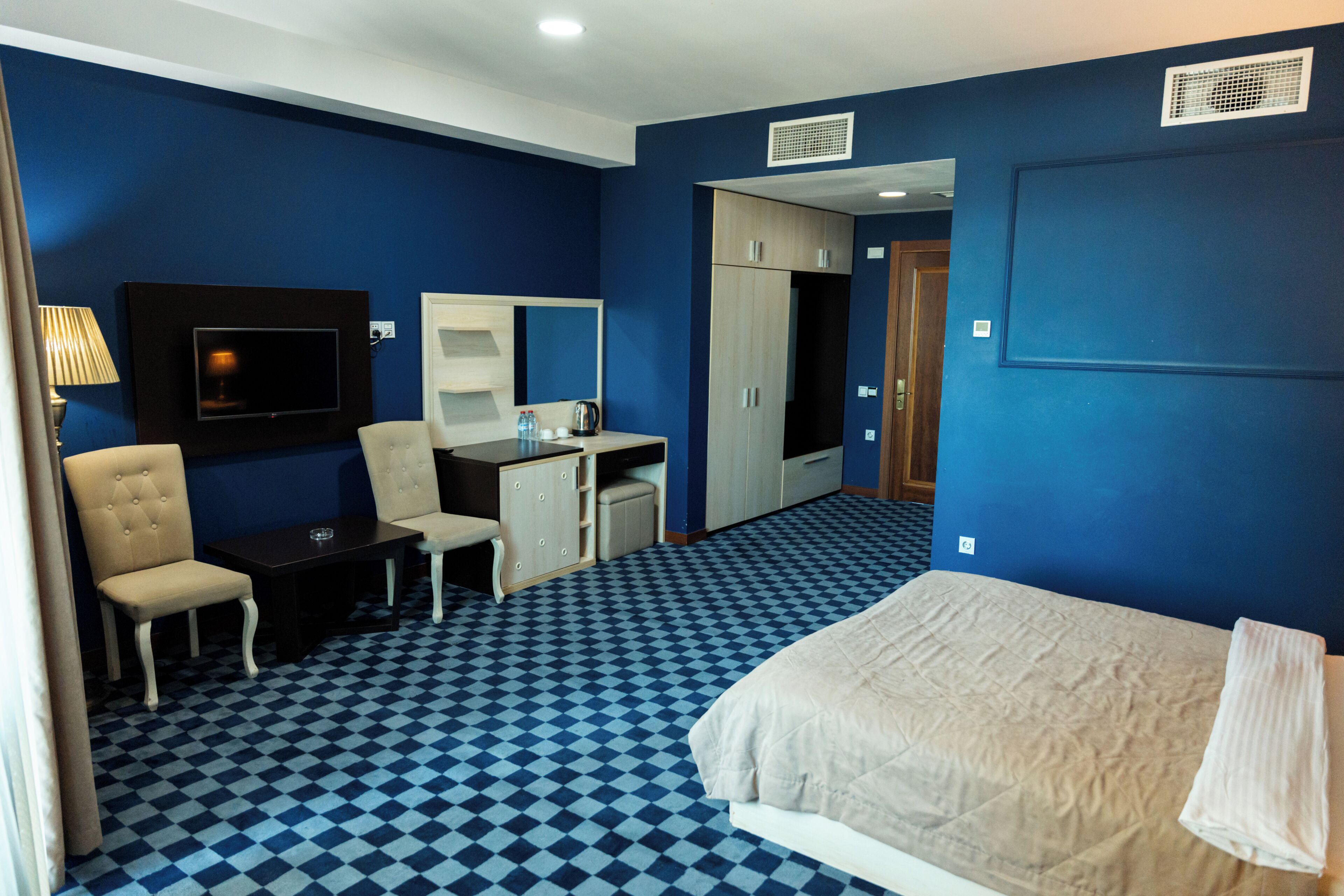 Ein Hotelzimmer mit Schachbrettboden, einem Bett, einem Schreibtisch, einem Stuhl, einem Fernseher und einem Kleiderschrank.