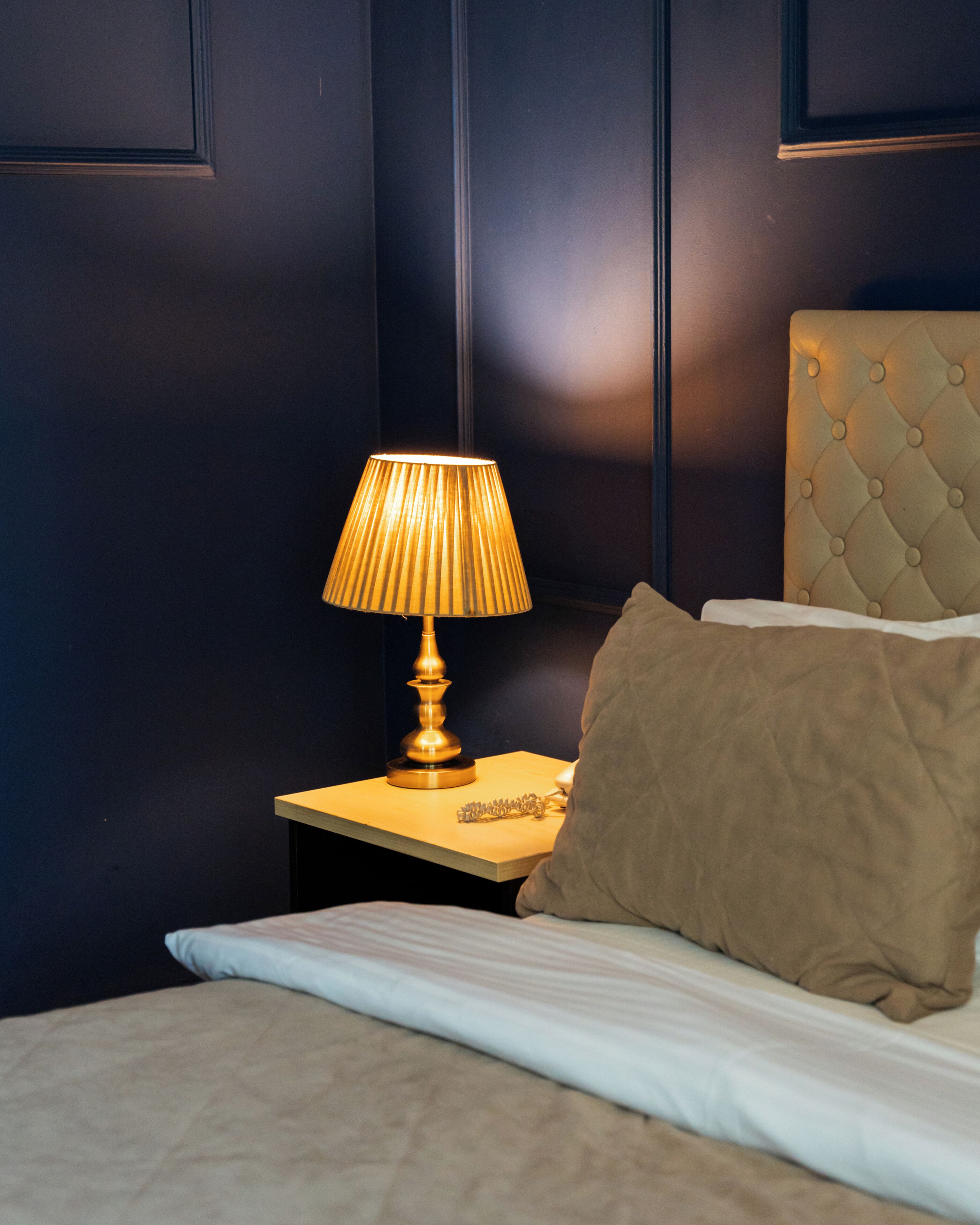 A neatly made bed with a white comforter and a brown pillow, a bedside lamp on a wooden nightstand, and a tufted headboard.
